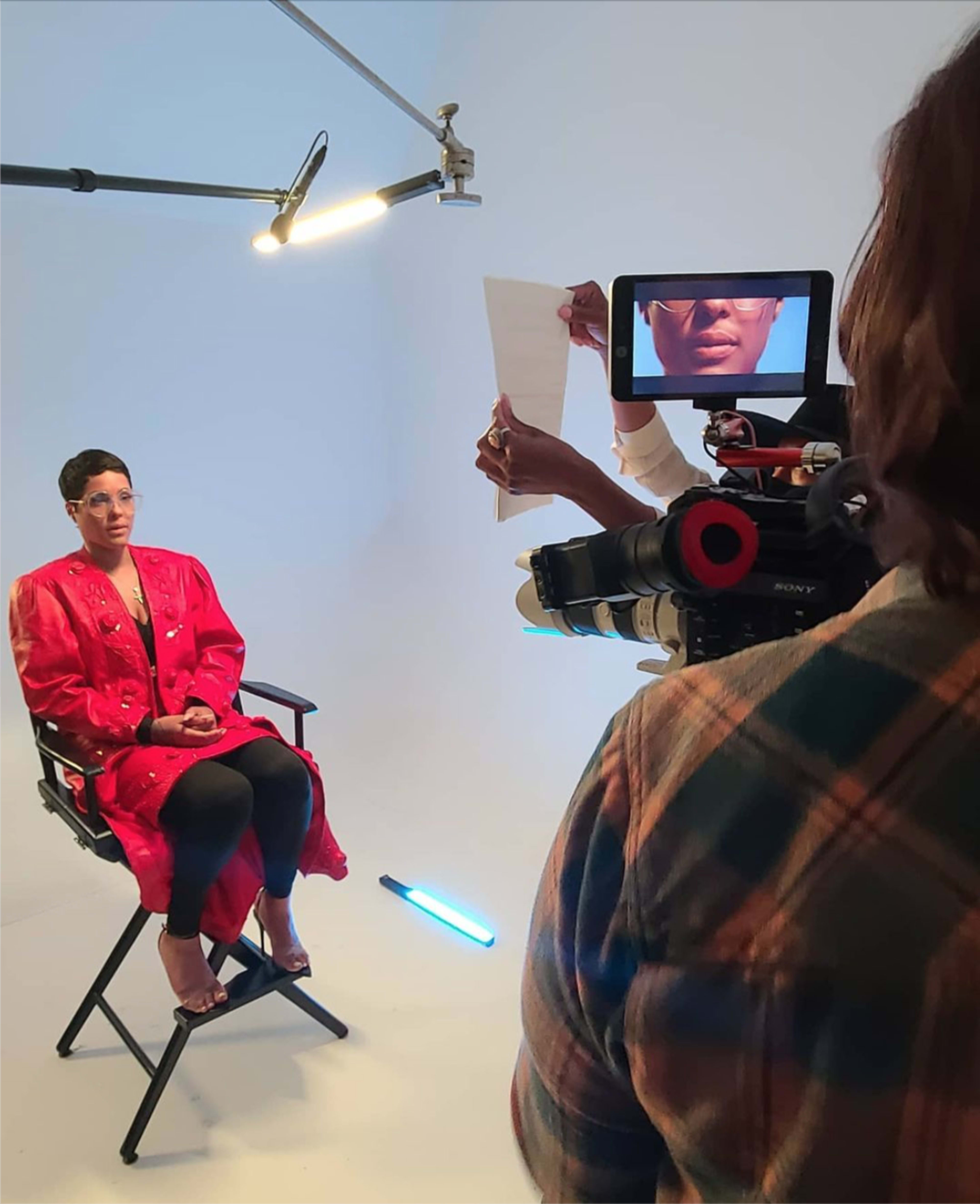 A woman sitting in a director's chair is being filmed in a studio, while another person holds a script in front of a camera monitor.