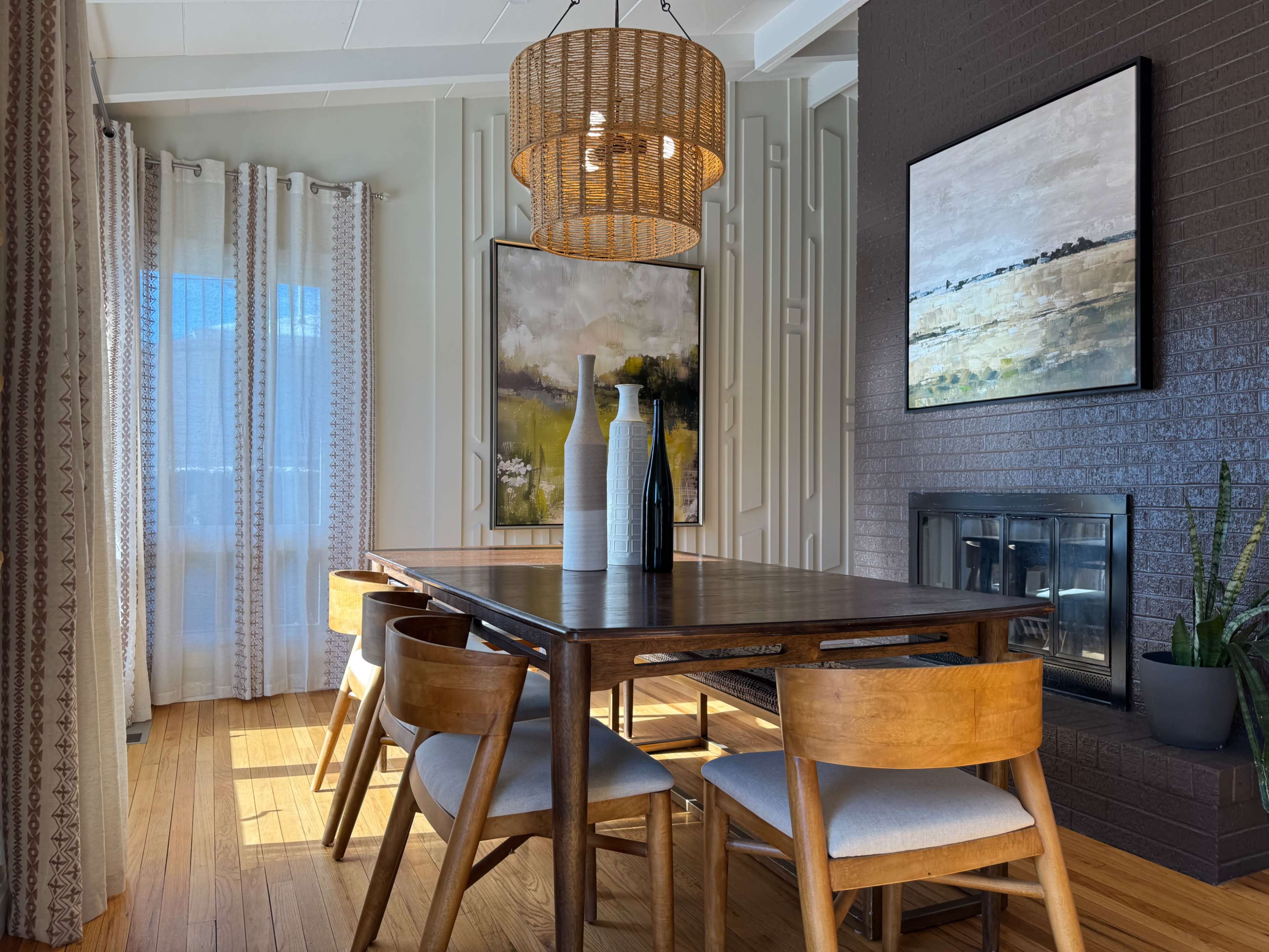 A dining area features a wooden table surrounded by four chairs, with a woven pendant light overhead, a large painting on the wall, and a fireplace in the background.