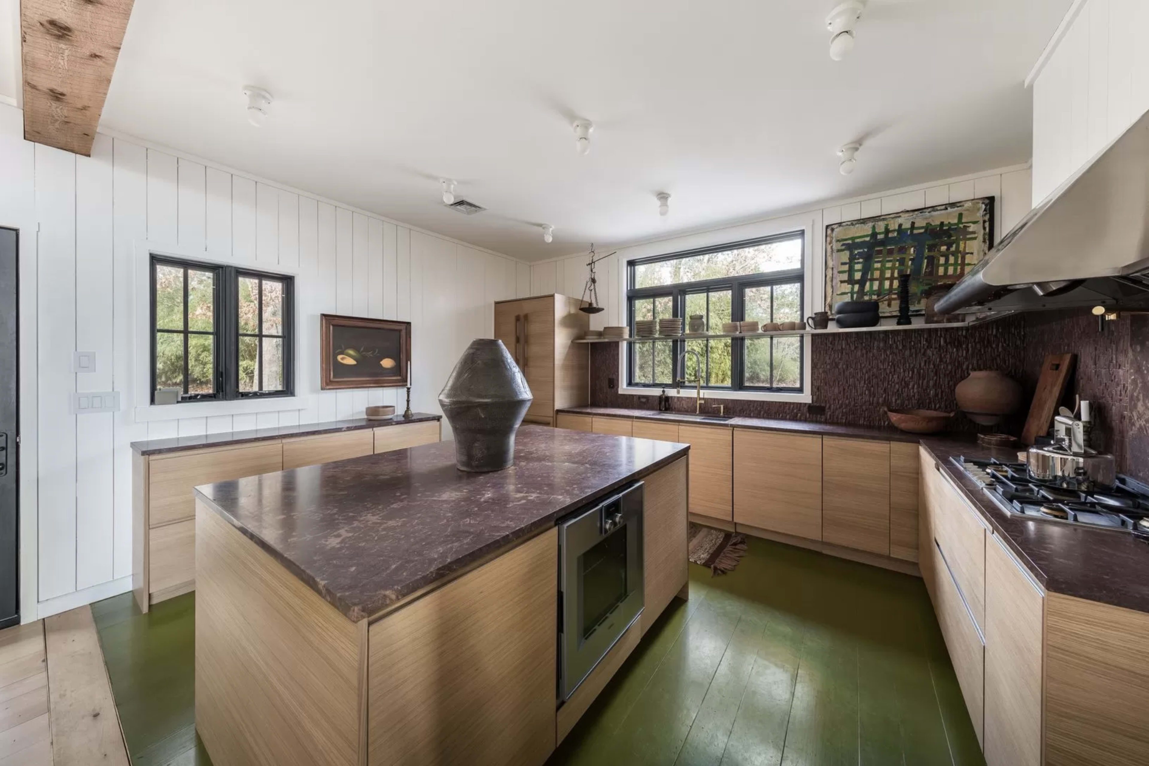 The kitchen features a central island with a dark countertop, wooden cabinetry, and large windows allowing natural light.