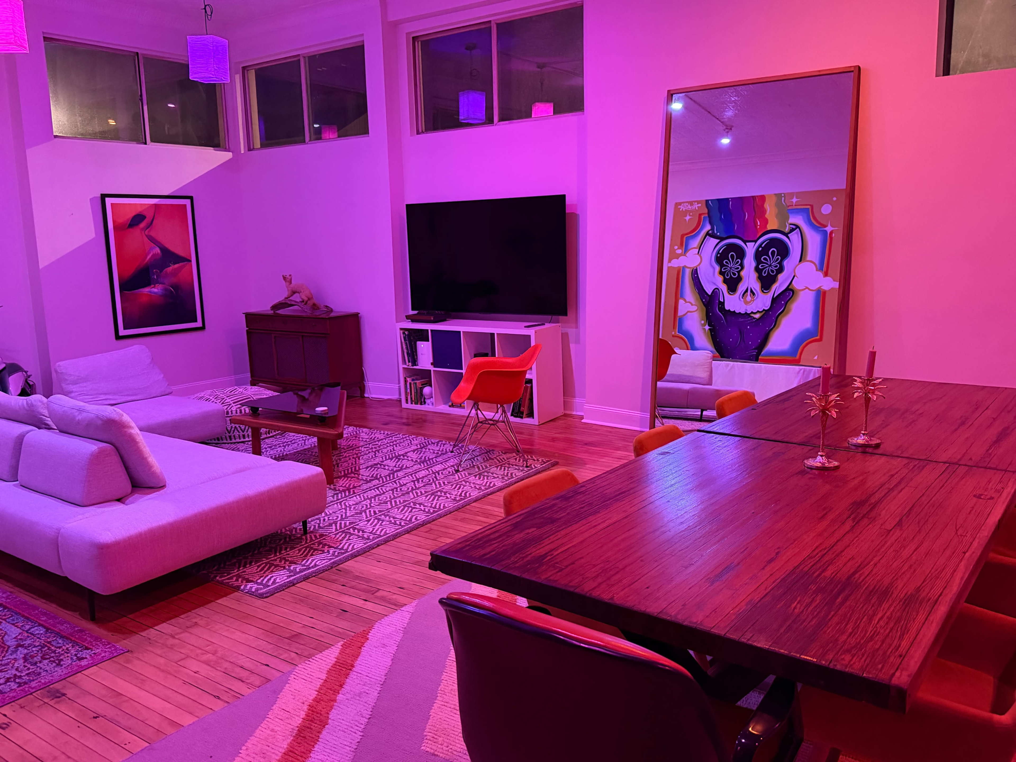 The image shows a modern living space with a pink-lit atmosphere, featuring a sectional sofa, a large television, a dining table, and artwork on the walls.