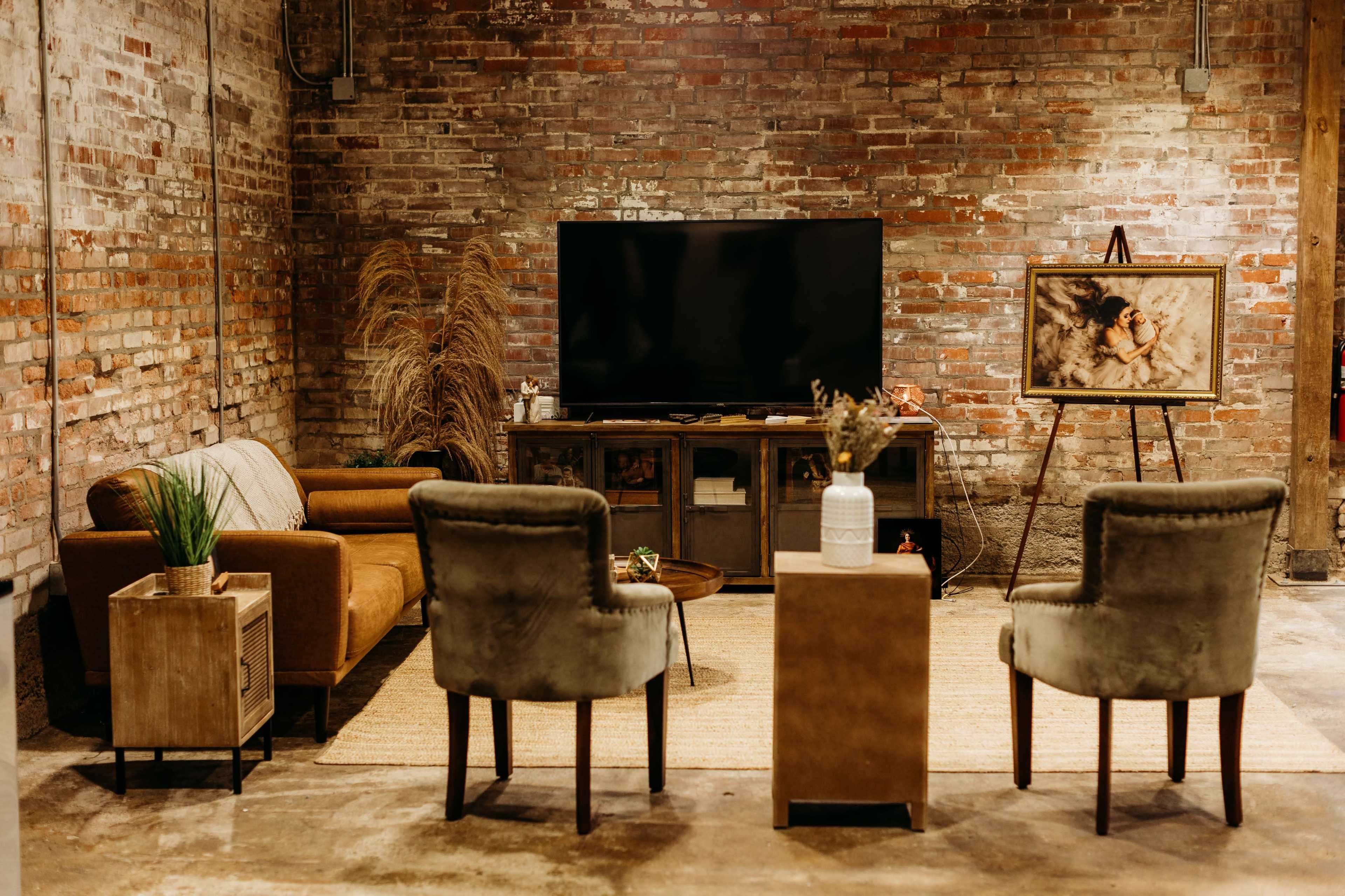 A cozy living room features a television against a brick wall, with an arrangement of furniture including a brown sofa, two upholstered chairs, and a side table, alongside a framed artwork on an easel.