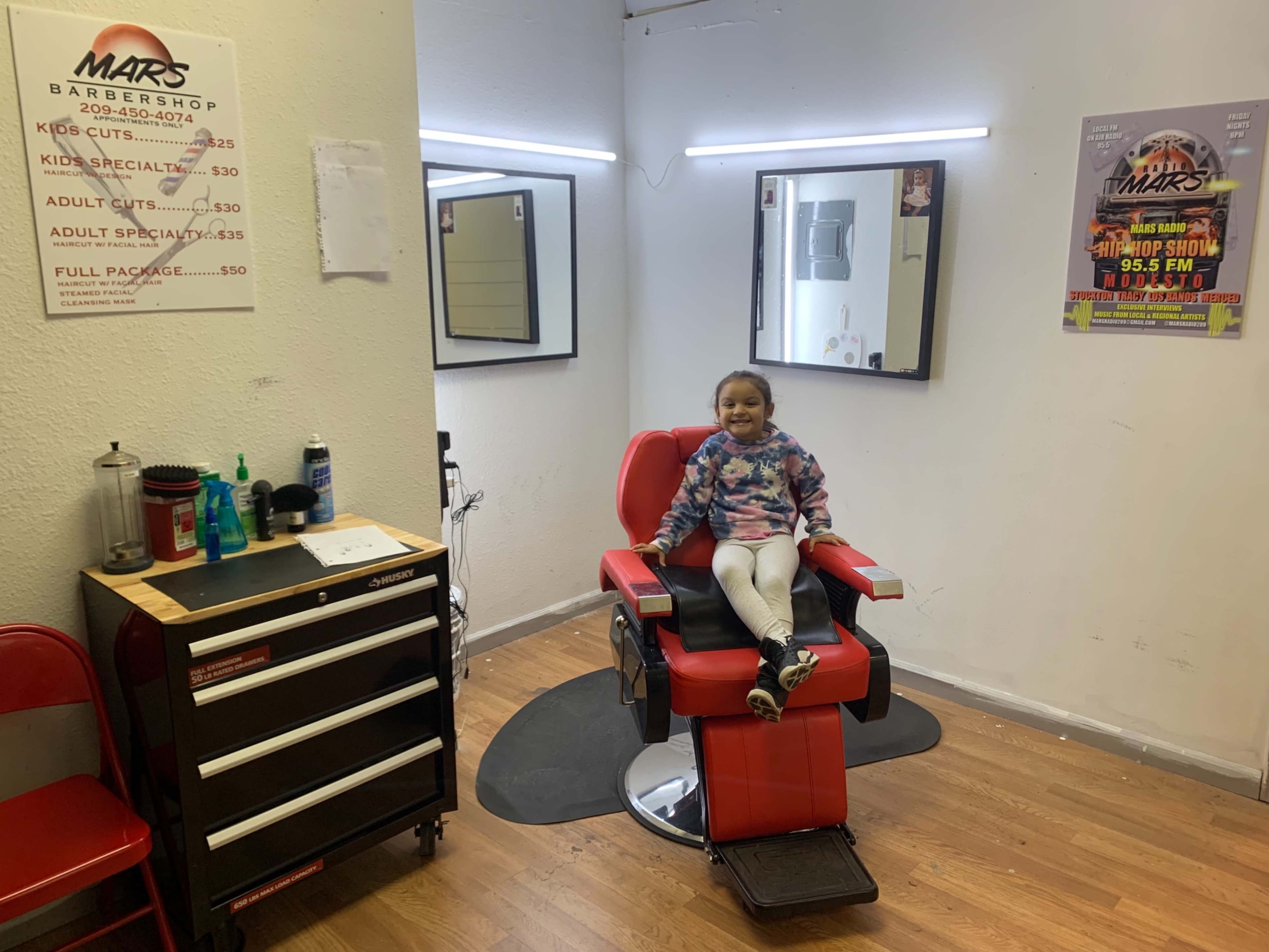 A young girl sits on a red barber chair in a barber shop, surrounded by mirrors and a tool cart.