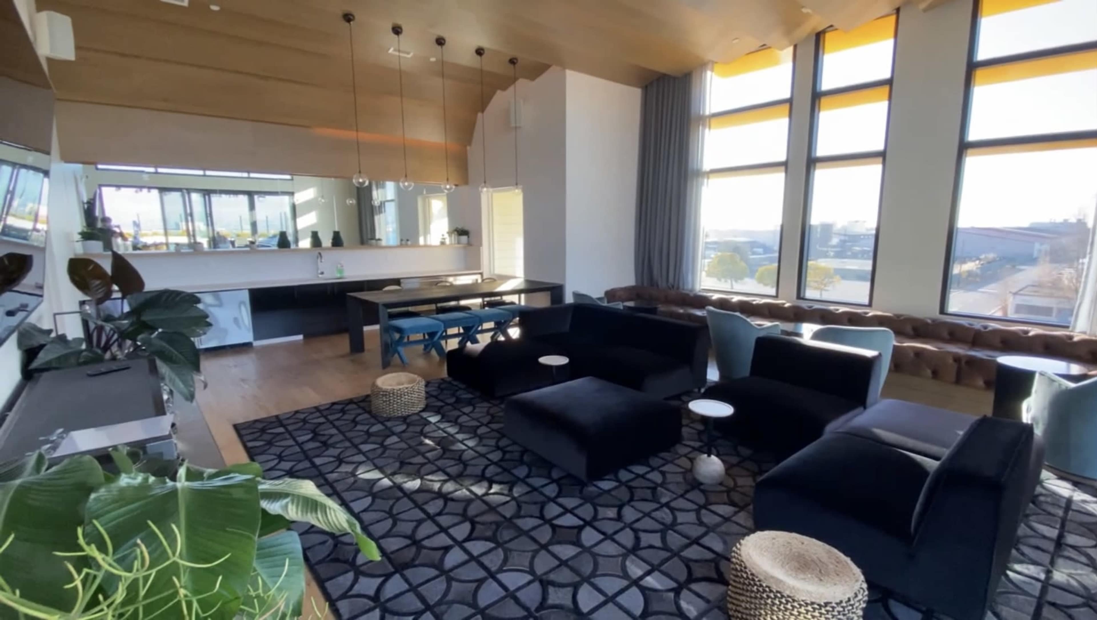 A modern lounge area featuring dark furniture, large windows with natural light, and a patterned rug.