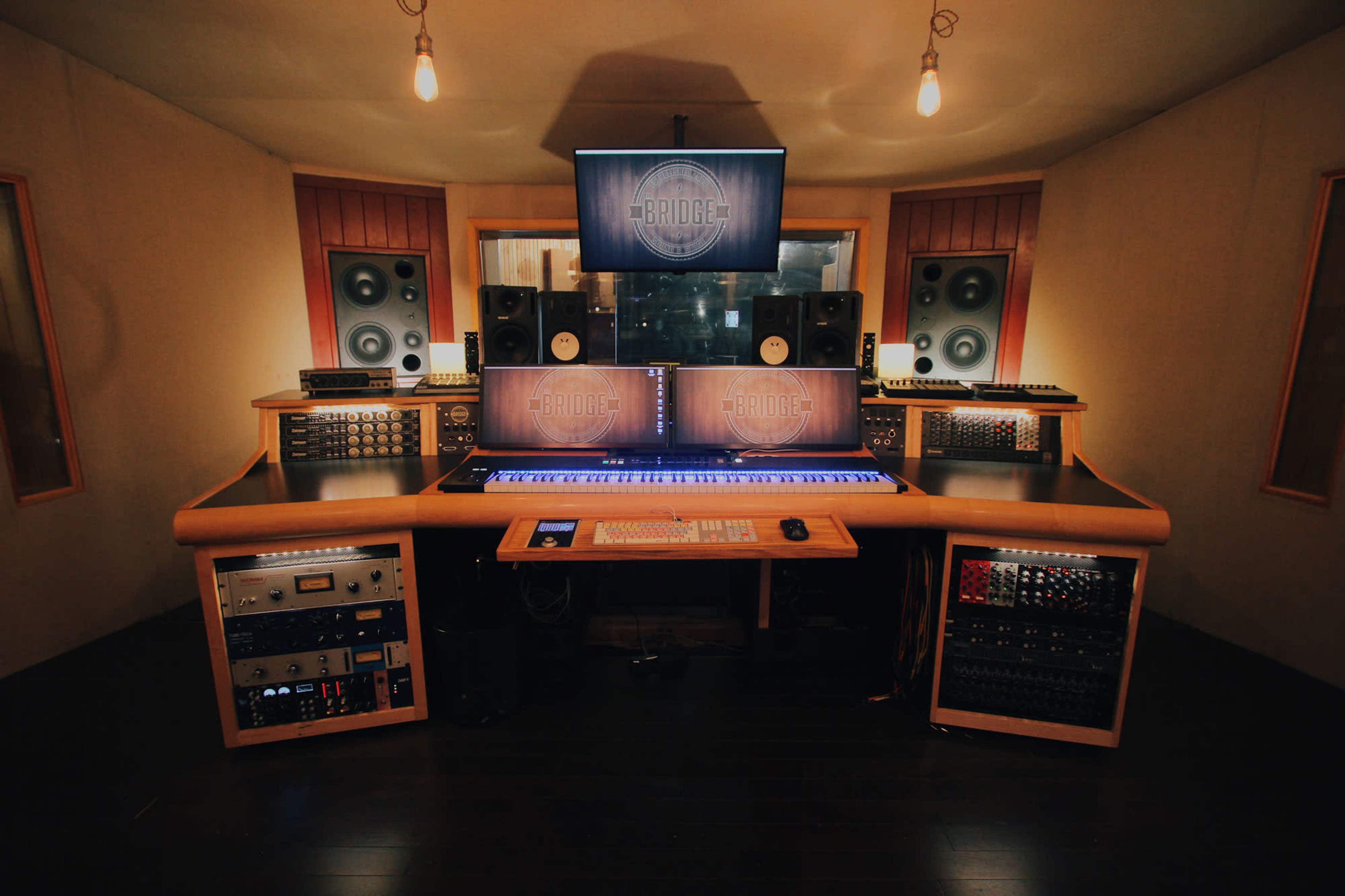 The image shows a professional music recording studio with a central mixing console, screens, and various audio equipment arranged on wall-mounted shelves.
