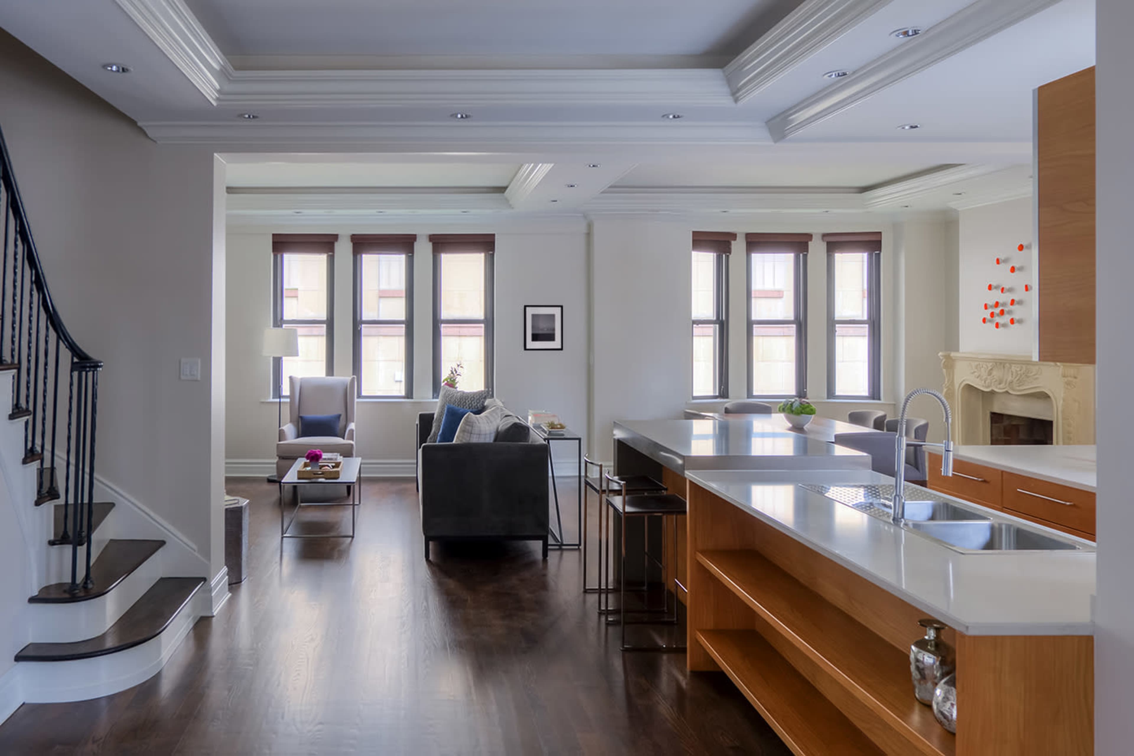A modern open-concept living space features a kitchen with wooden cabinetry, a dining area with stools, and a living room with large windows and comfortable seating.