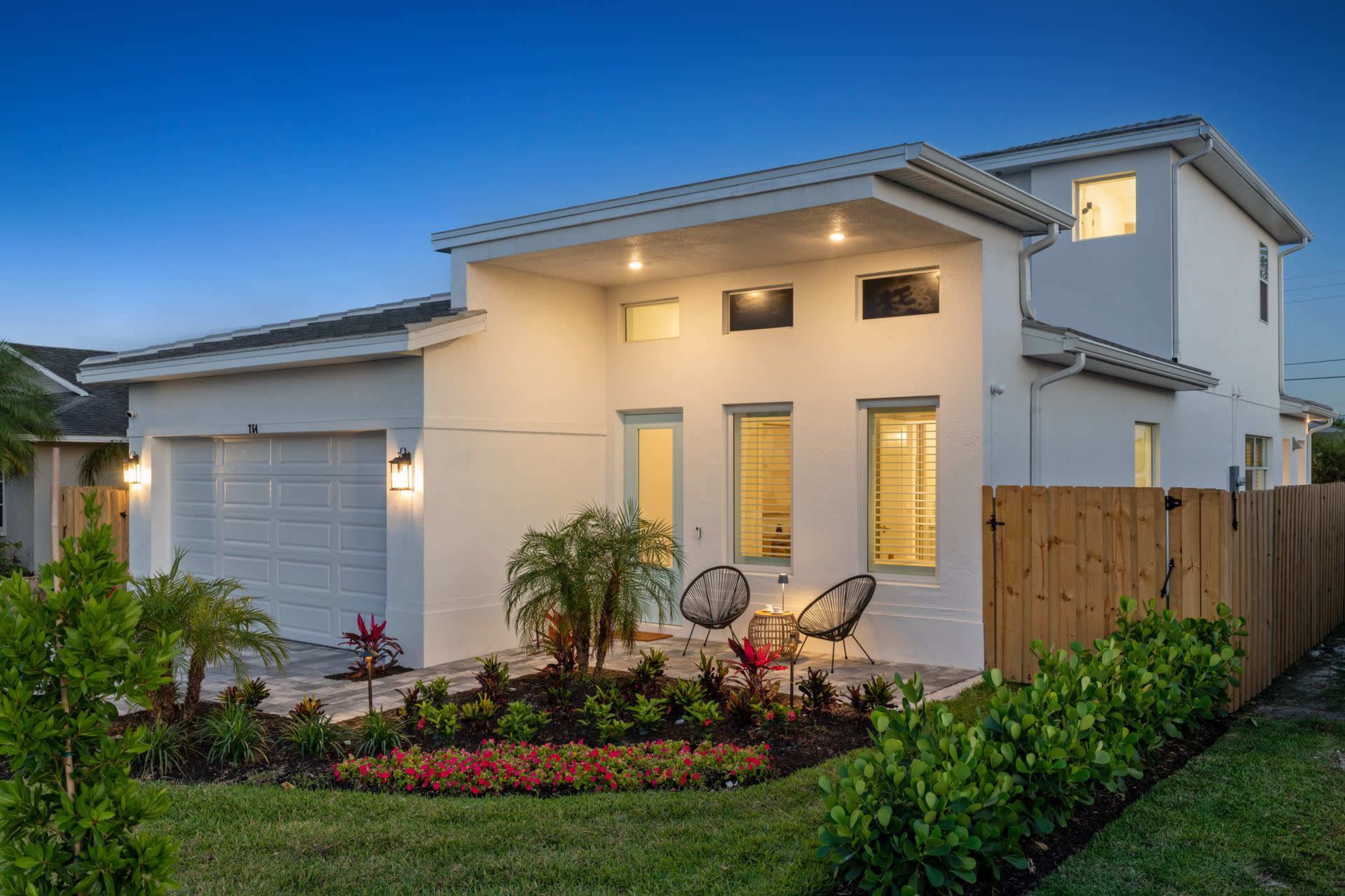 A modern, two-story white house with a well-lit exterior, a landscaped front yard, and a paved entrance featuring two chairs.