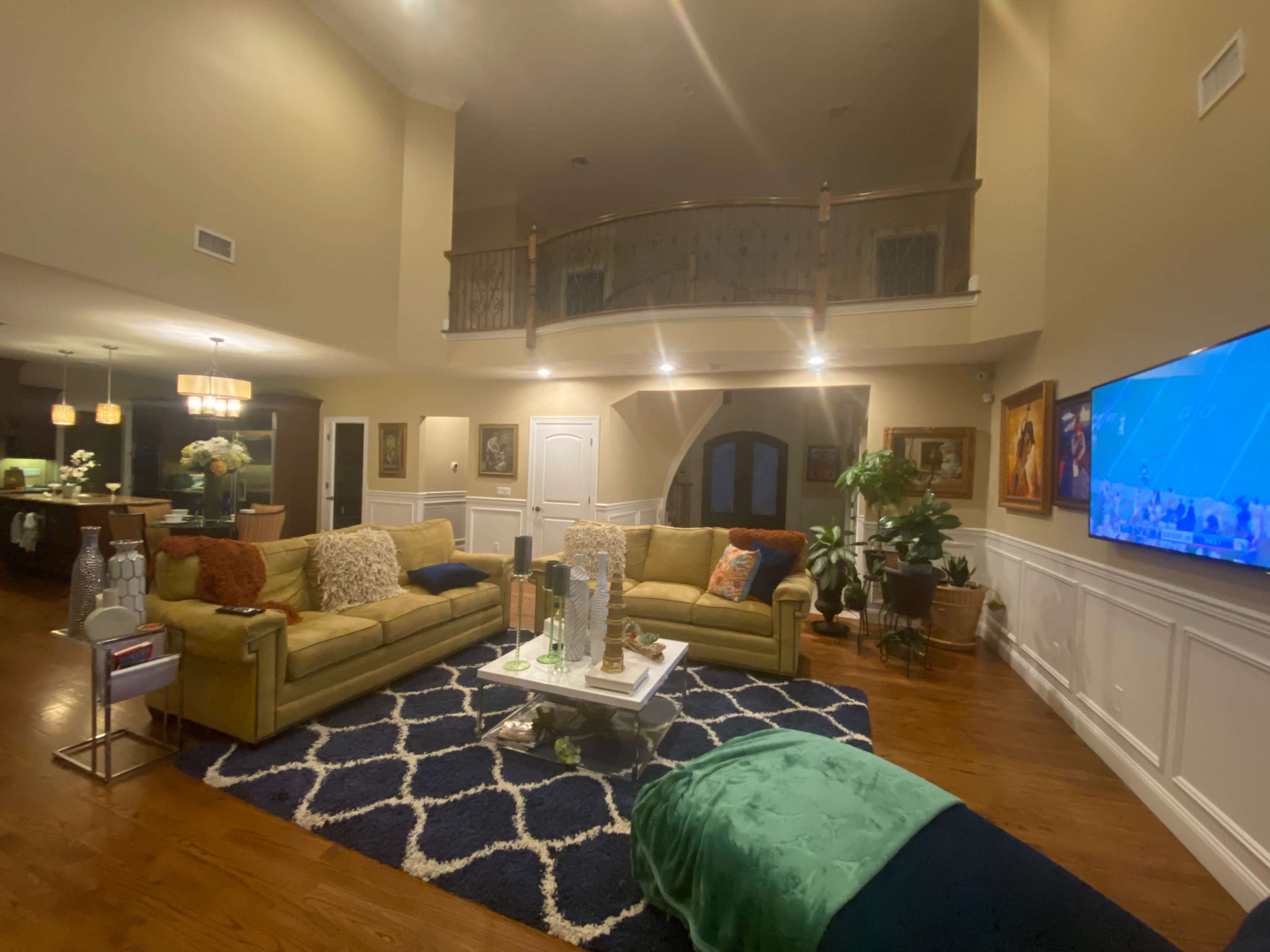 A spacious living room with two yellow sofas facing a coffee table, surrounded by artwork and decorative plants, and featuring a staircase leading to a balcony.