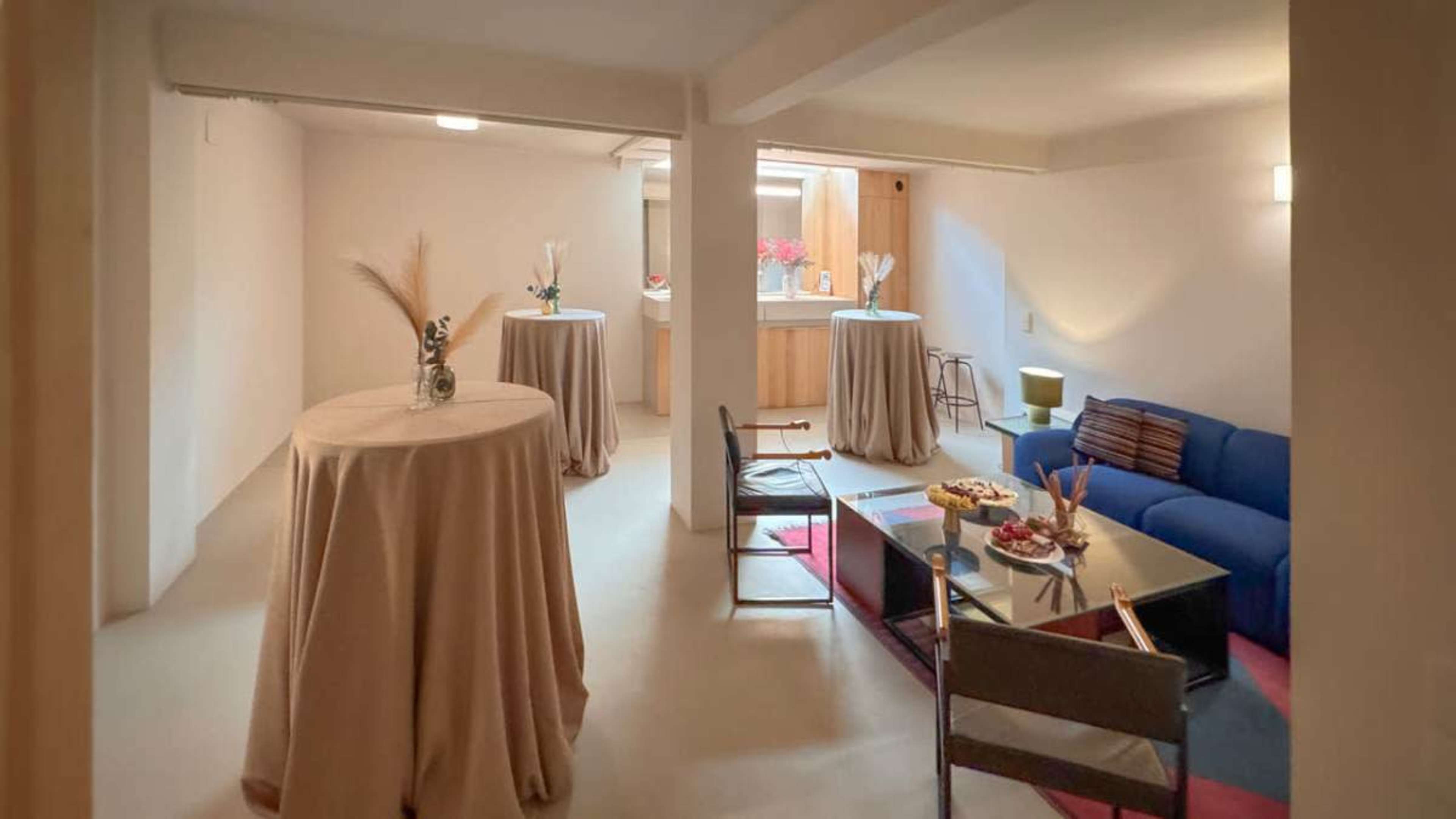 The image depicts a modern, minimalist living space with a blue sofa, a coffee table set with snacks, and several tall tables draped in beige cloth.