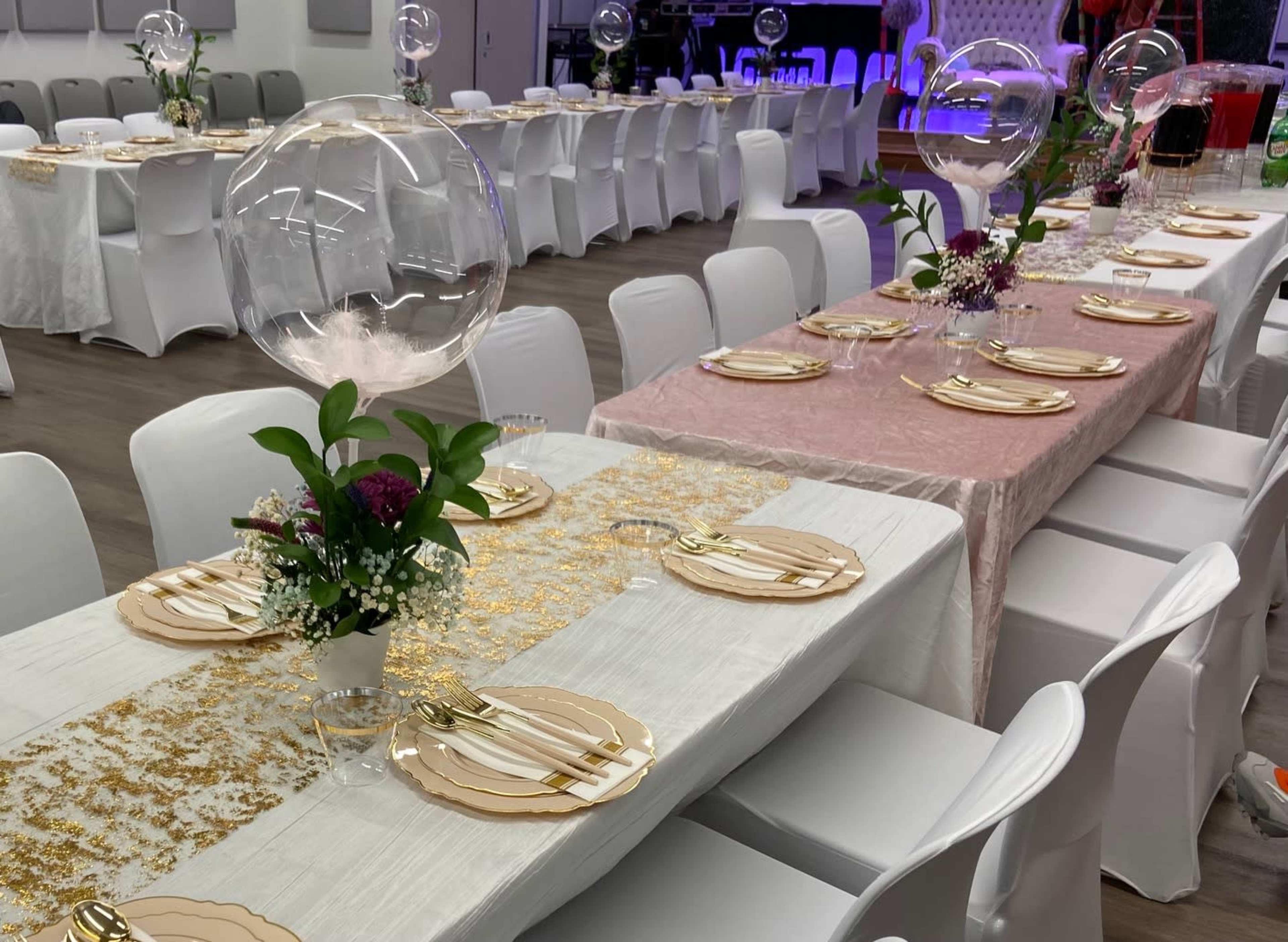 Two long banquet tables are set with elegant place settings, floral arrangements, and decorative glass orbs in a brightly lit event space.