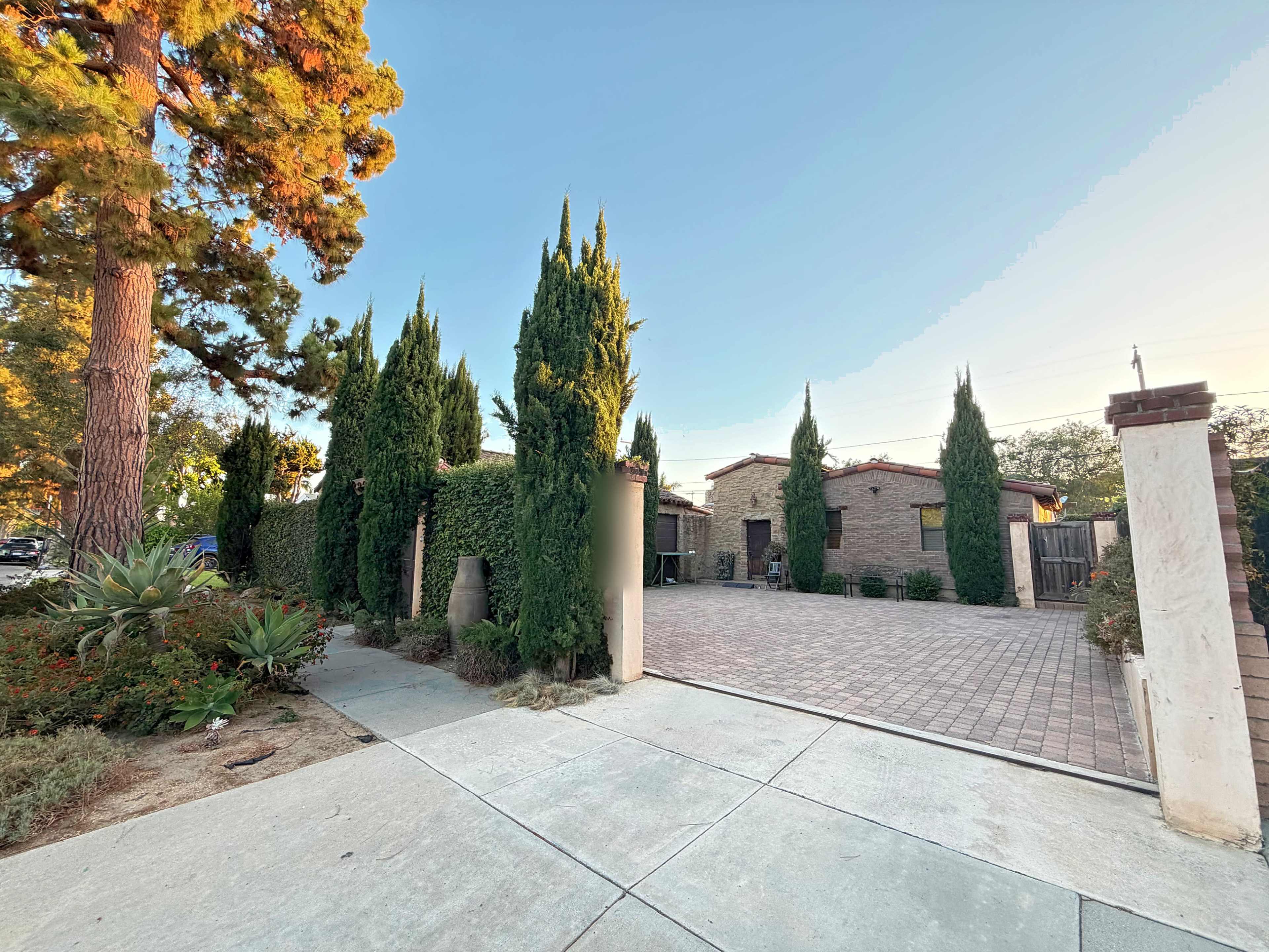 A pathway leads to a landscaped yard featuring tall greenery and a brick-paved driveway in front of a residential building.
