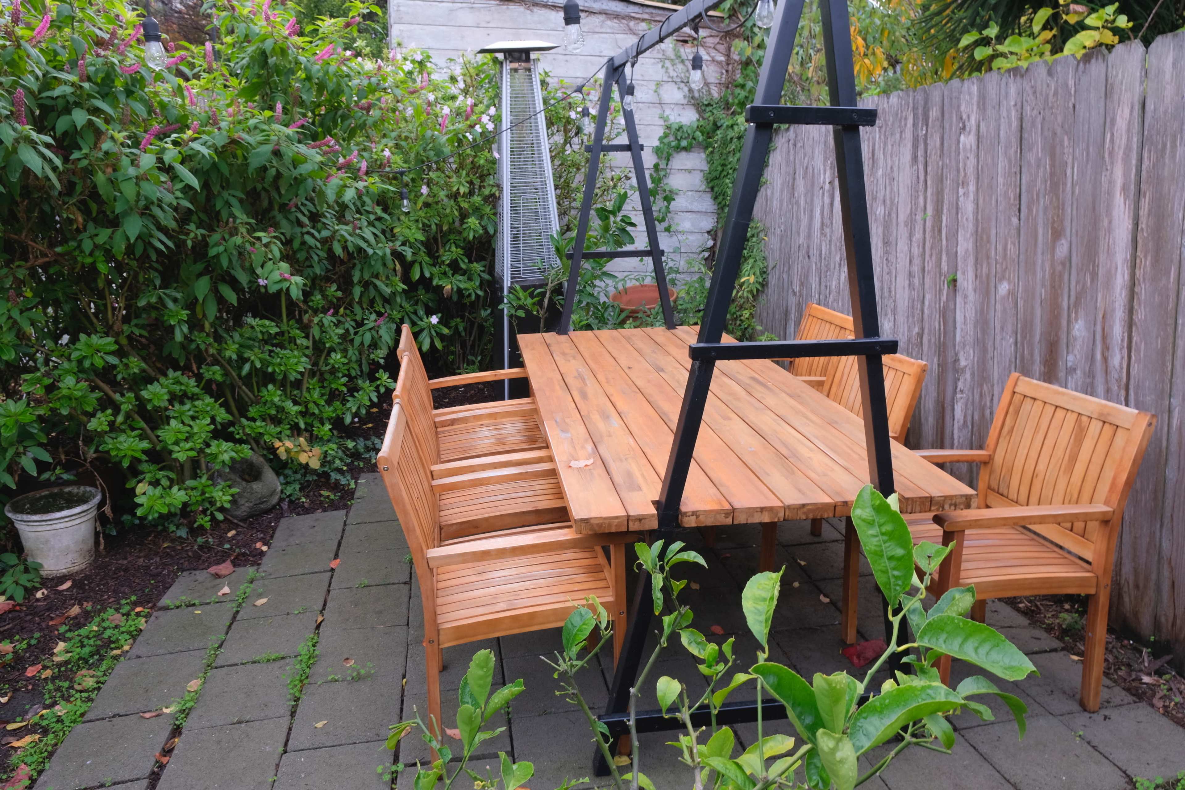 A wooden patio table with chairs is set in a small outdoor space surrounded by greenery and a wooden fence.