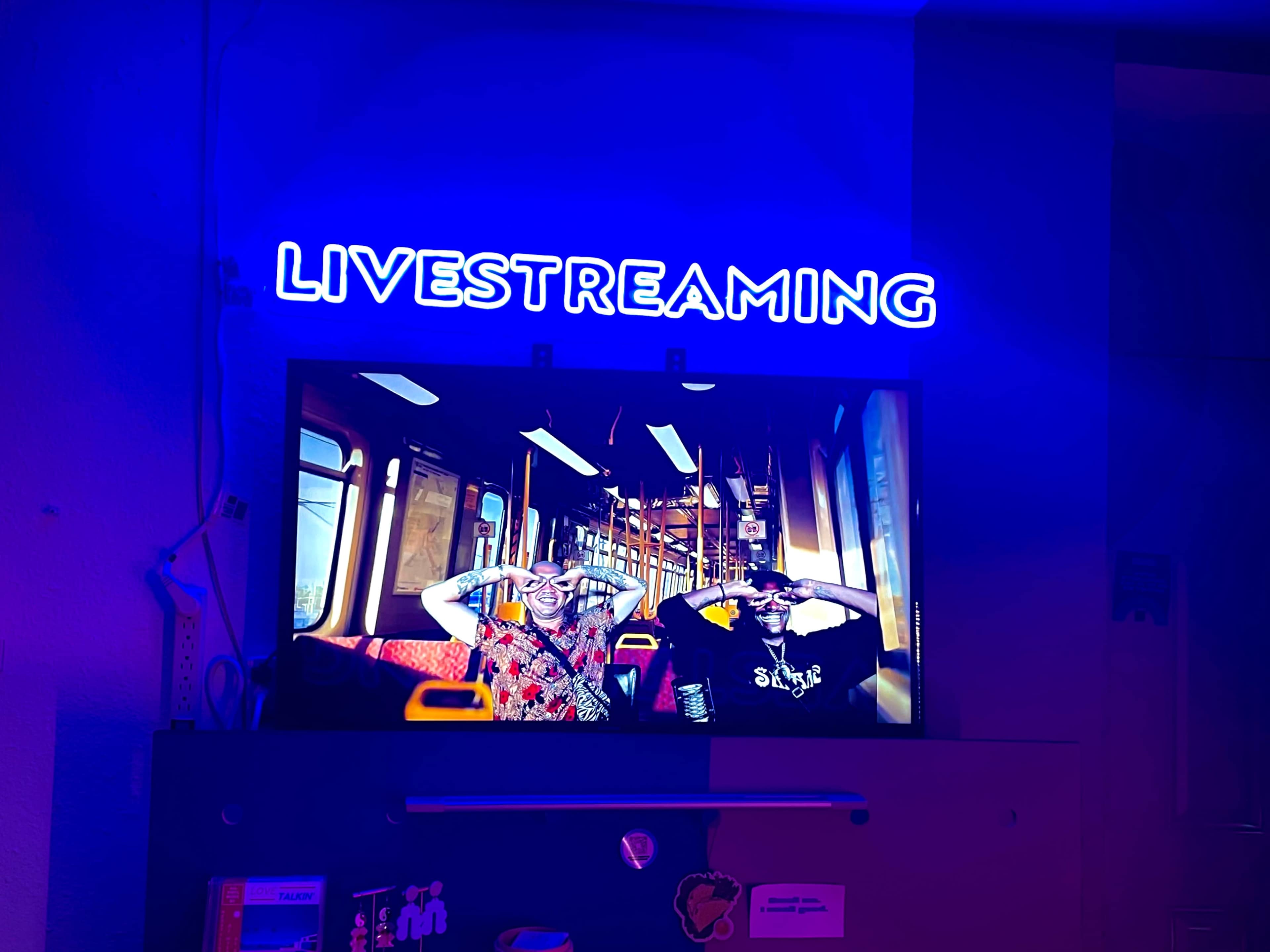 A neon sign reads "LIVESTREAMING" above a television displaying two people in a train car.