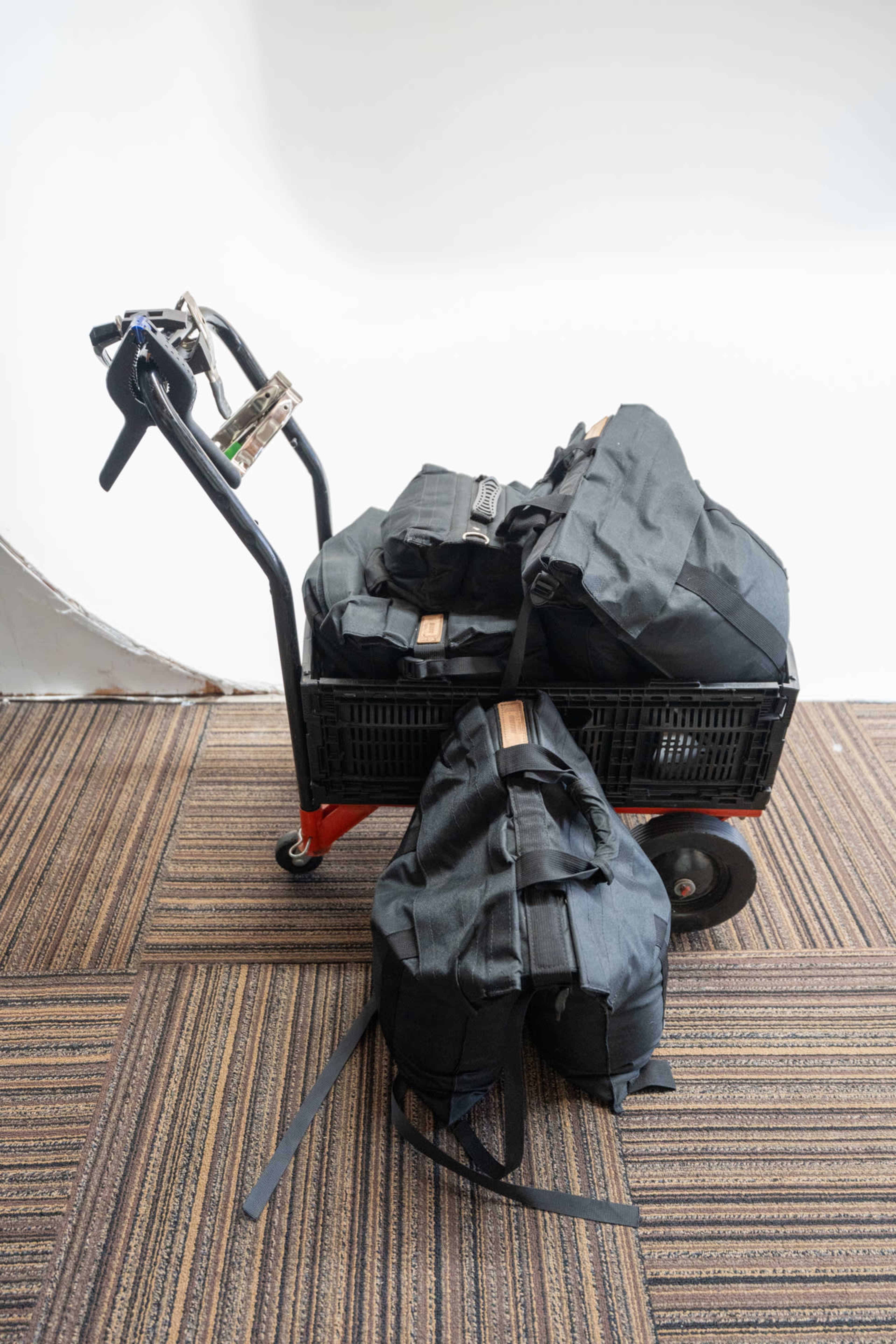 A black cart holds several large black backpacks on a striped carpet surface.