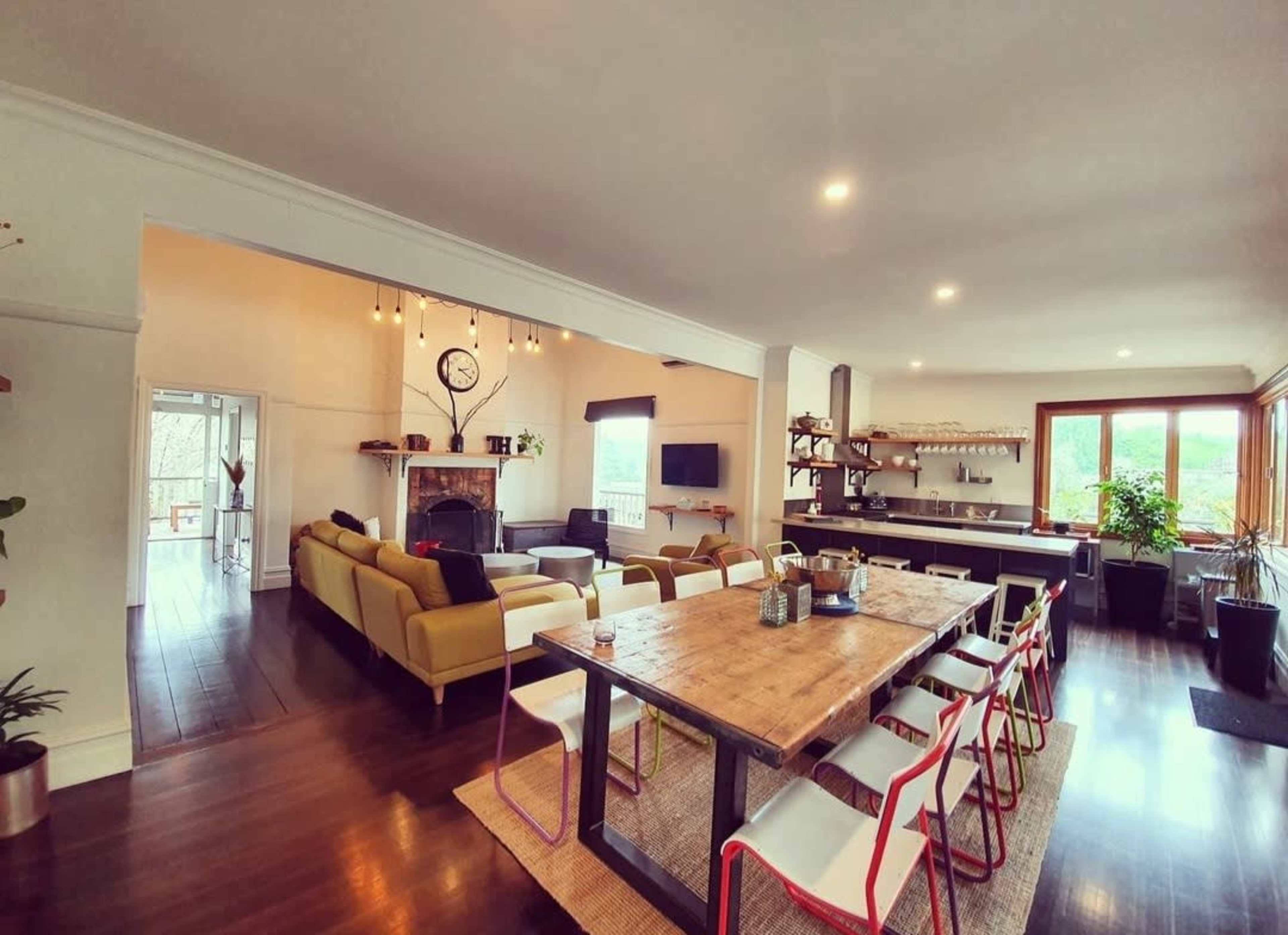 An open-concept living and dining area featuring a long wooden table with colorful chairs, a cozy seating arrangement, and a modern kitchen in the background.