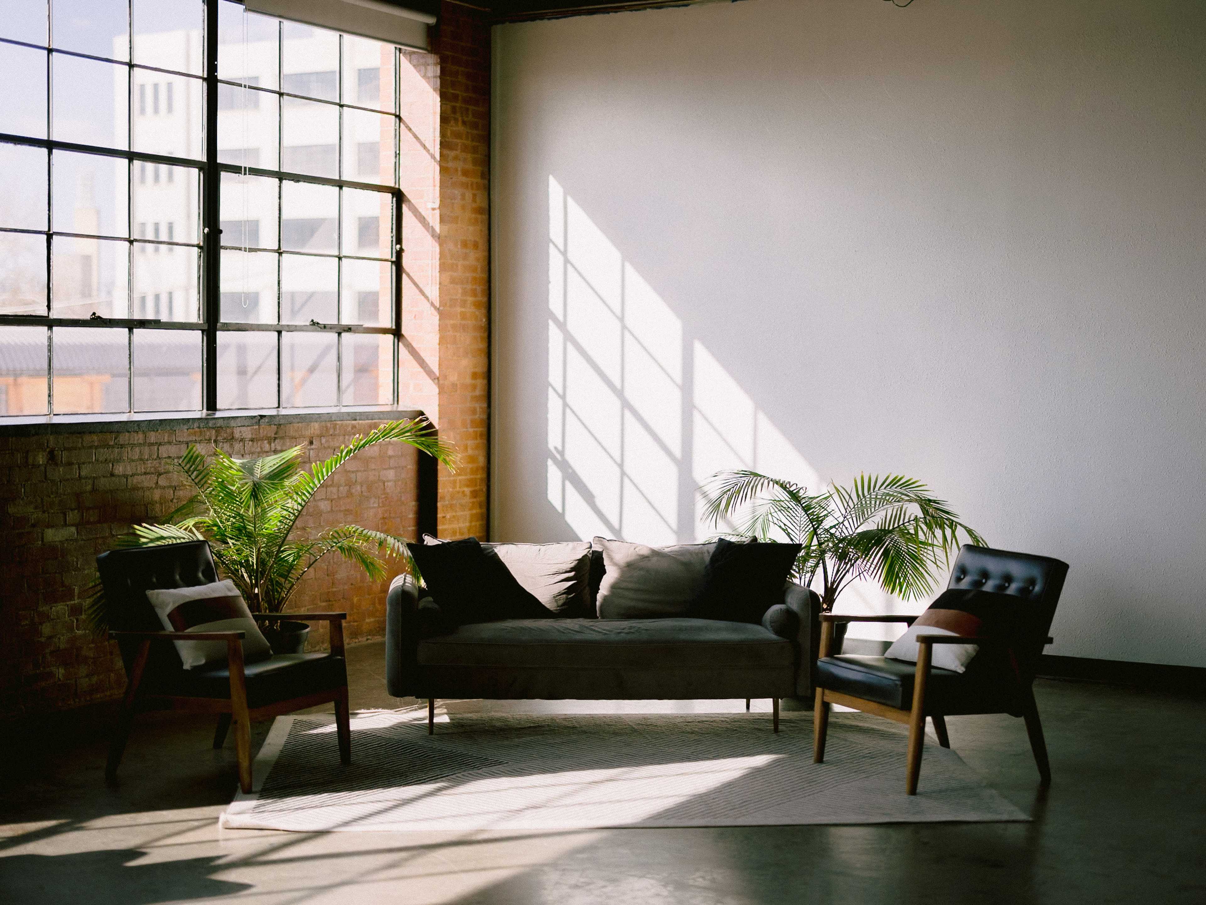 The image shows a modern seating area with a gray sofa and two black chairs, illuminated by sunlight streaming through large windows.