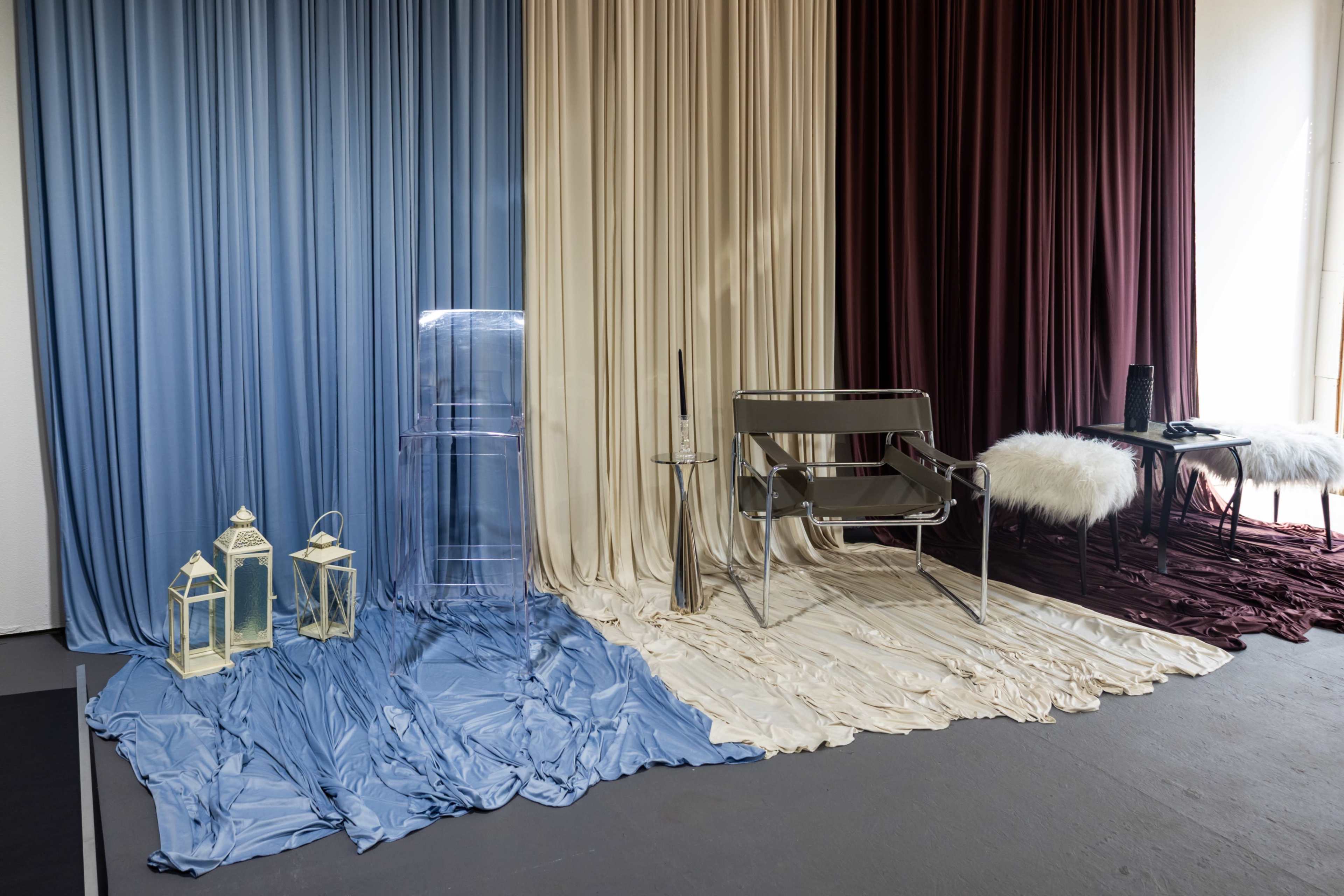 The image features a room divided into three sections with flowing fabric drapes in blue, creamy white, and dark red, accompanied by various furniture pieces and decorative lanterns.