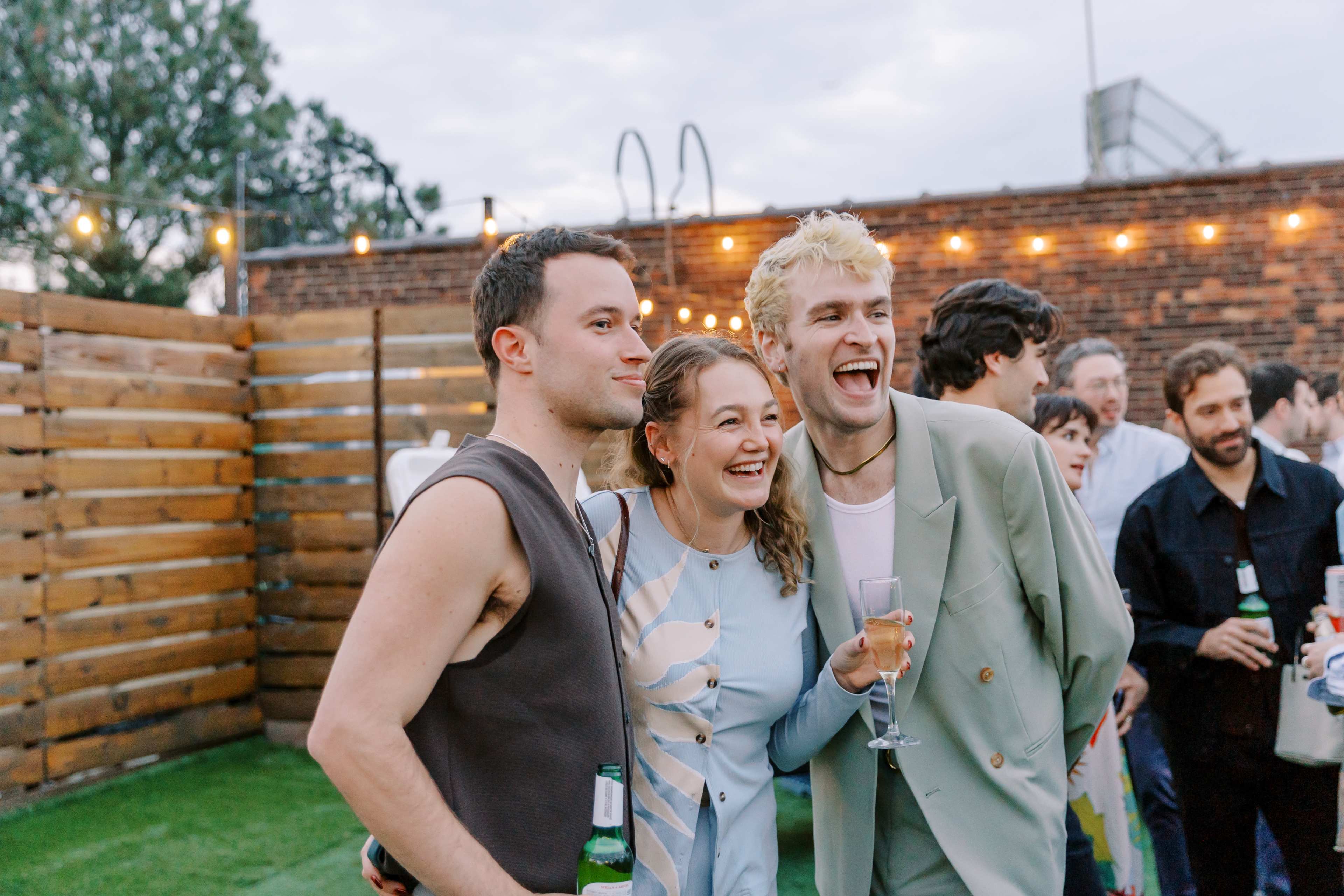 A group of three people smiles while posing together at a rooftop gathering with lights strung above.