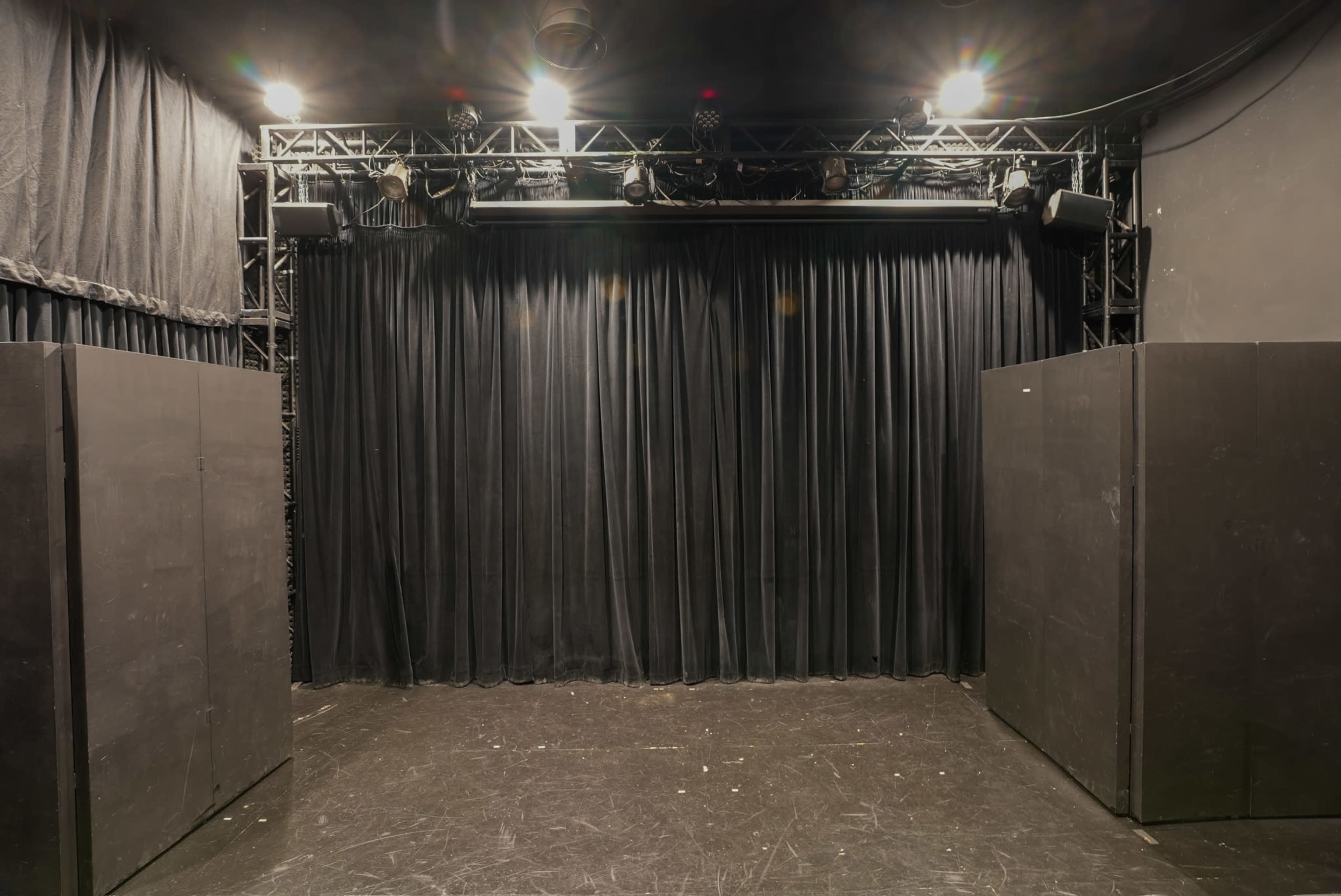 An empty stage with black curtains and lighting fixtures above it, surrounded by dark walls.
