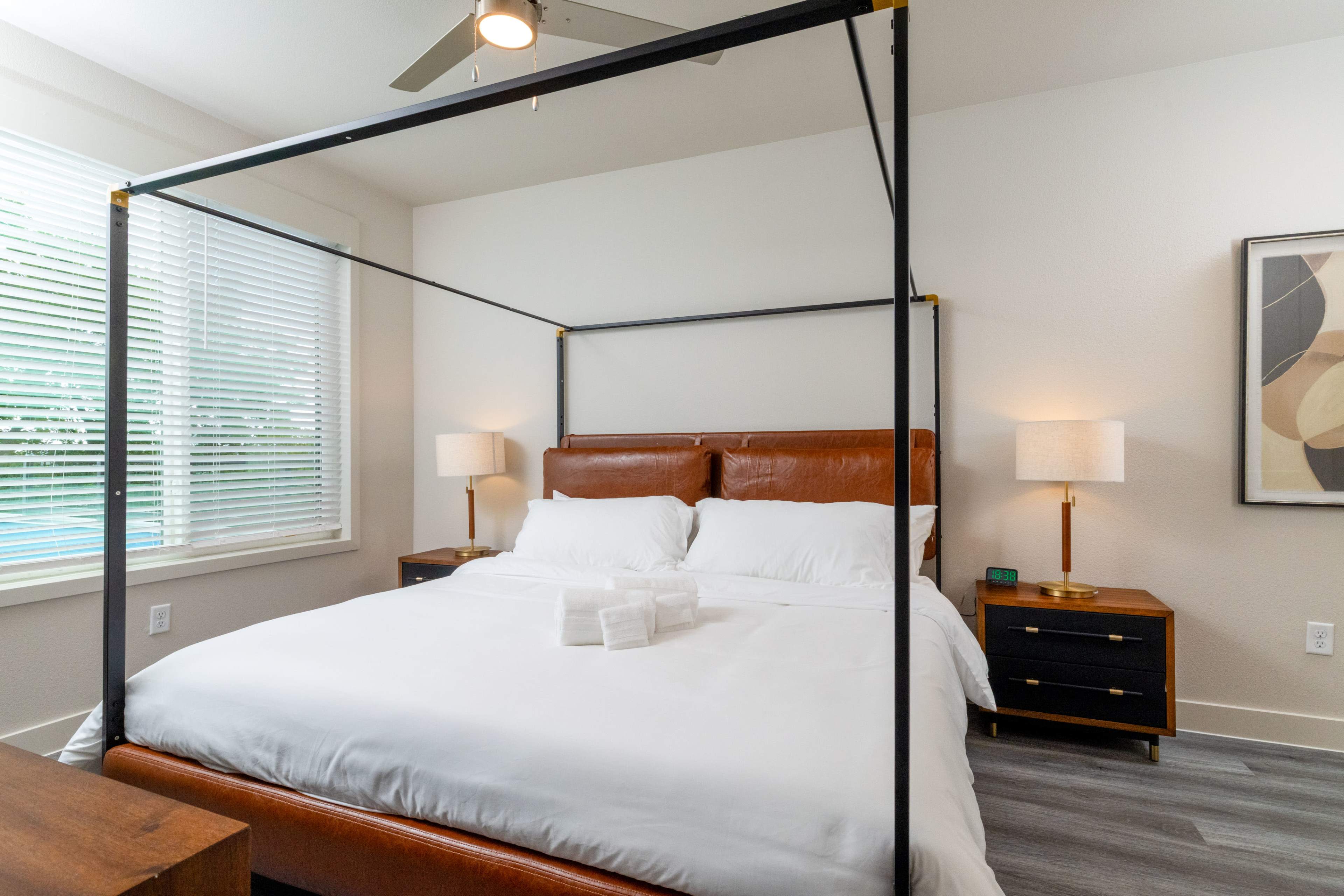The image shows a modern bedroom with a four-poster bed, white linens, and wooden nightstands on either side.
