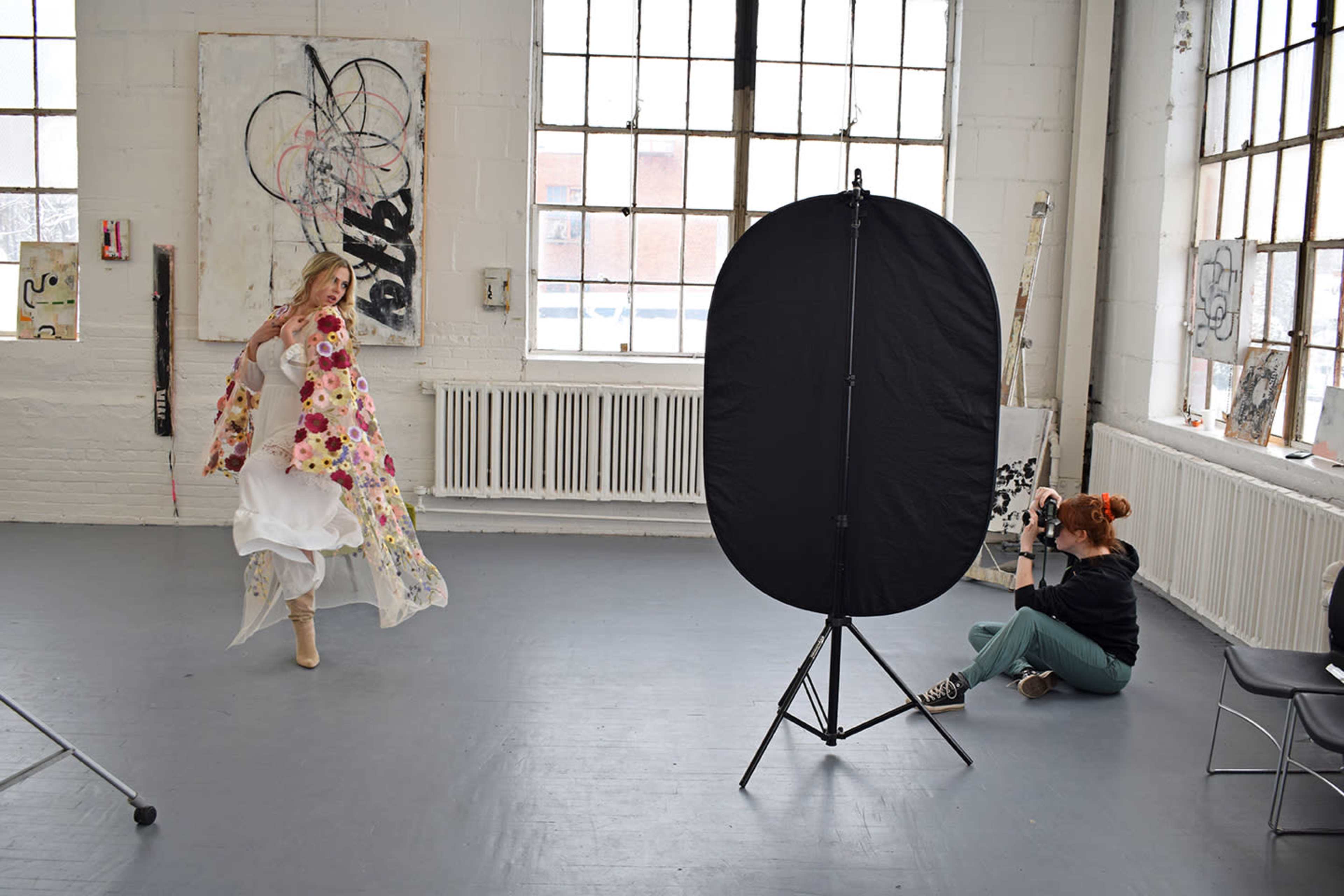 A woman in a floral wrap dances in a brightly lit studio while a photographer captures her image using a reflector.