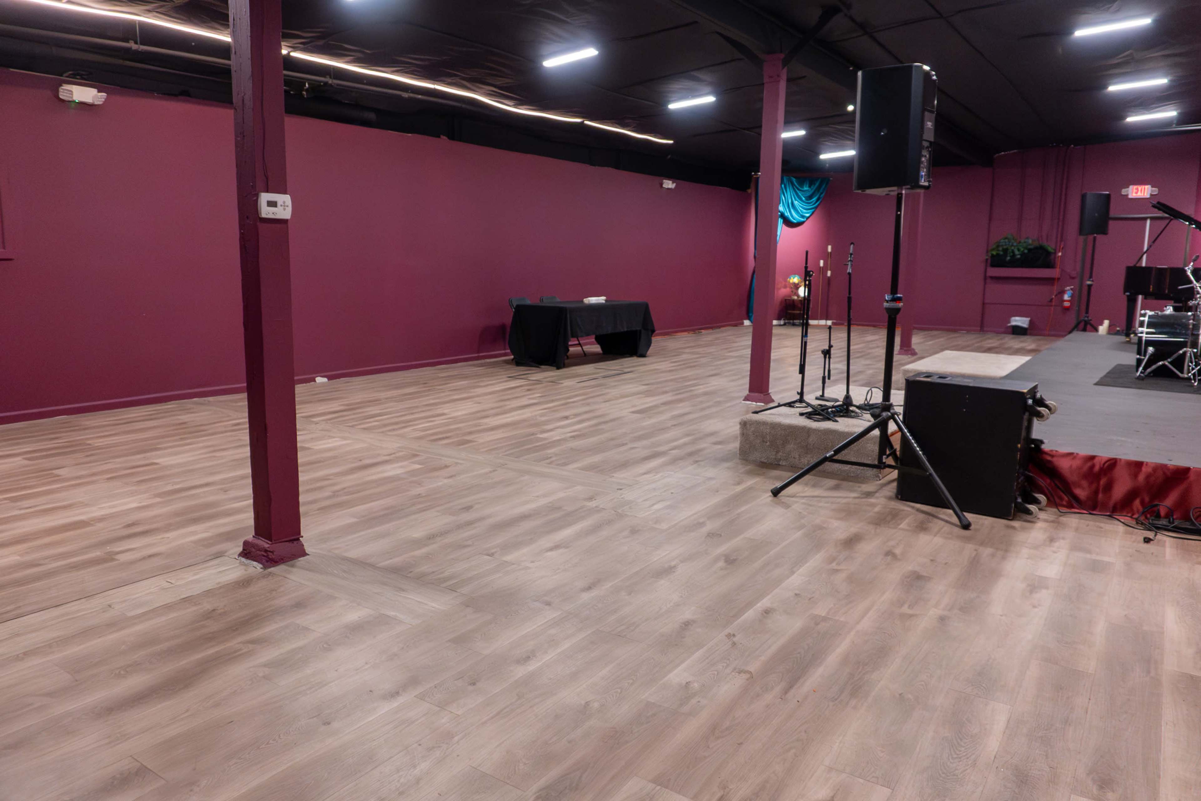 The image shows a spacious interior with wooden flooring, purple walls, a stage setup, a sound system, and a table along one side.
