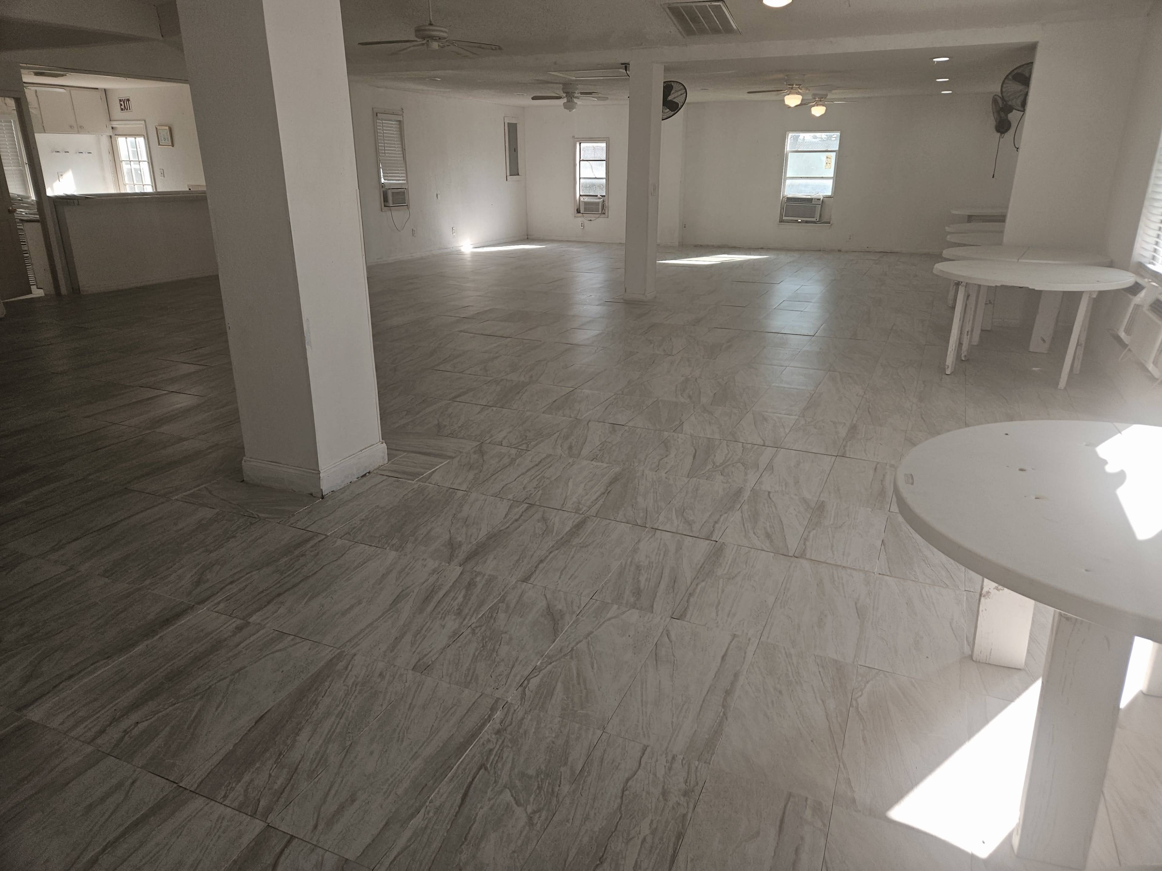 The image shows a spacious, empty room with tiled flooring and several ceiling fans, featuring a few round tables along the sides.