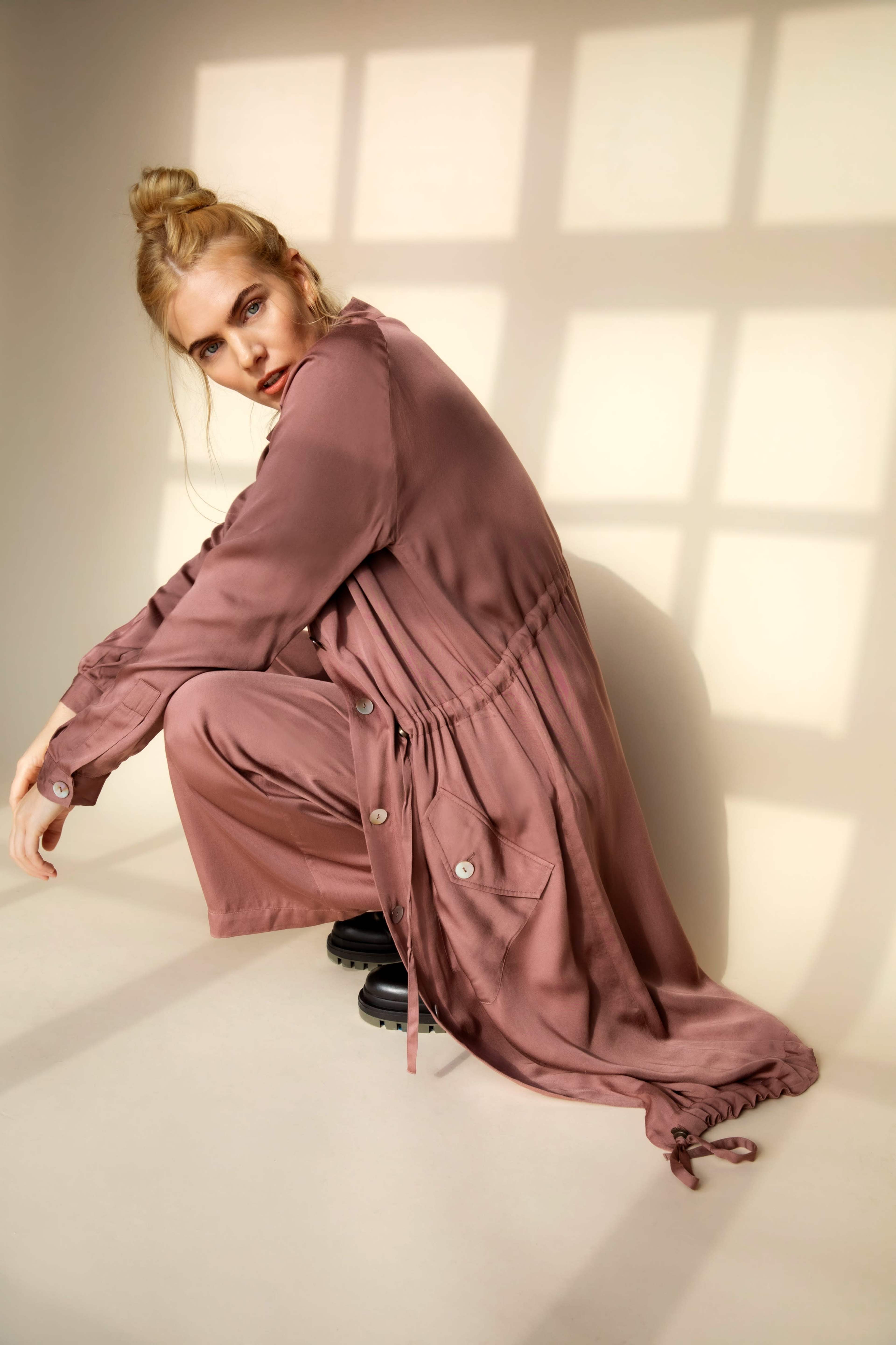 A person is sitting on the floor in a mauve outfit, leaning slightly forward with a serious expression, while shadows from a grid pattern are cast on the wall behind them.