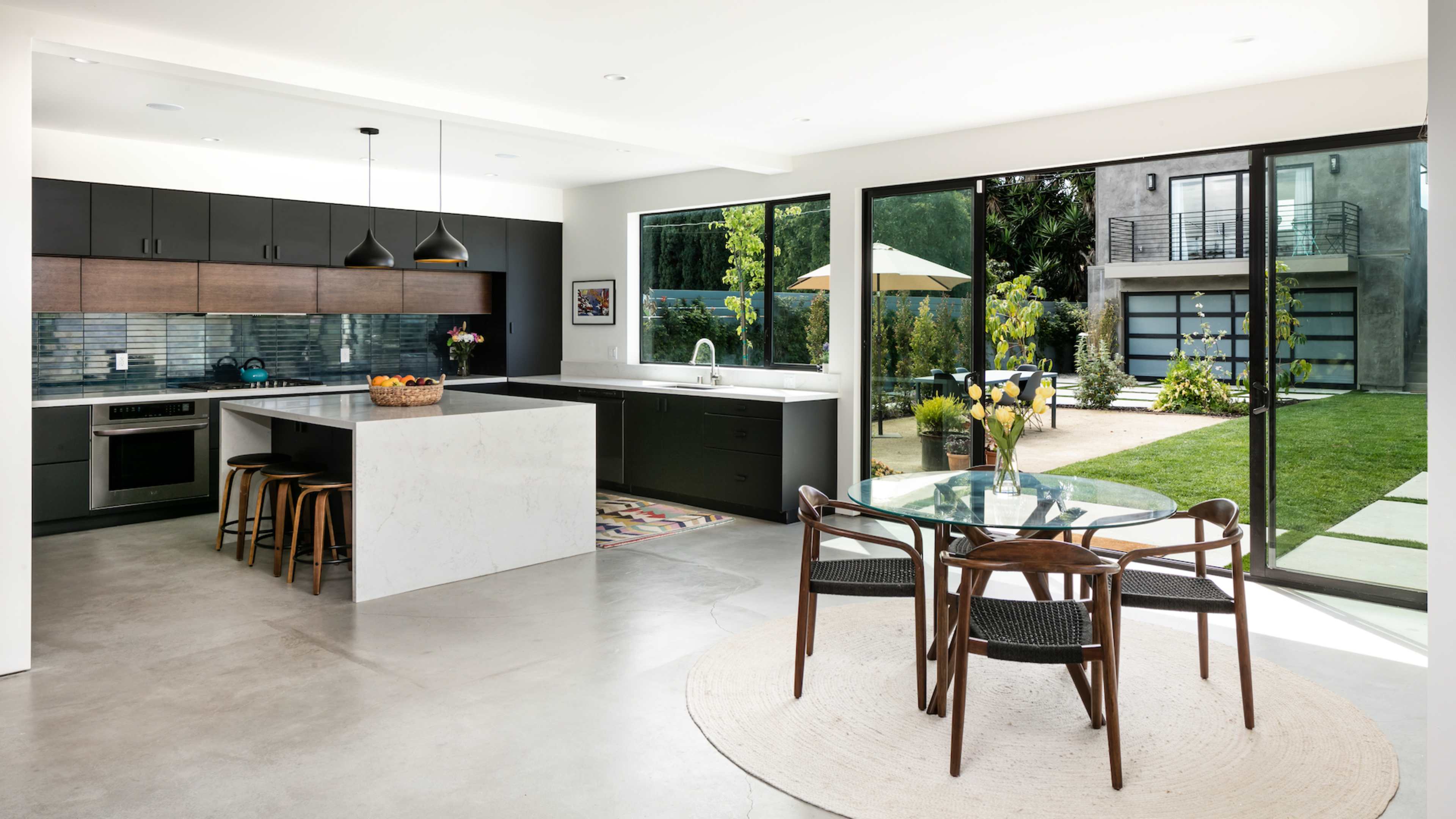 A modern kitchen and dining area with a large island, a round dining table, and glass doors leading to an outdoor space with a lawn and patio.