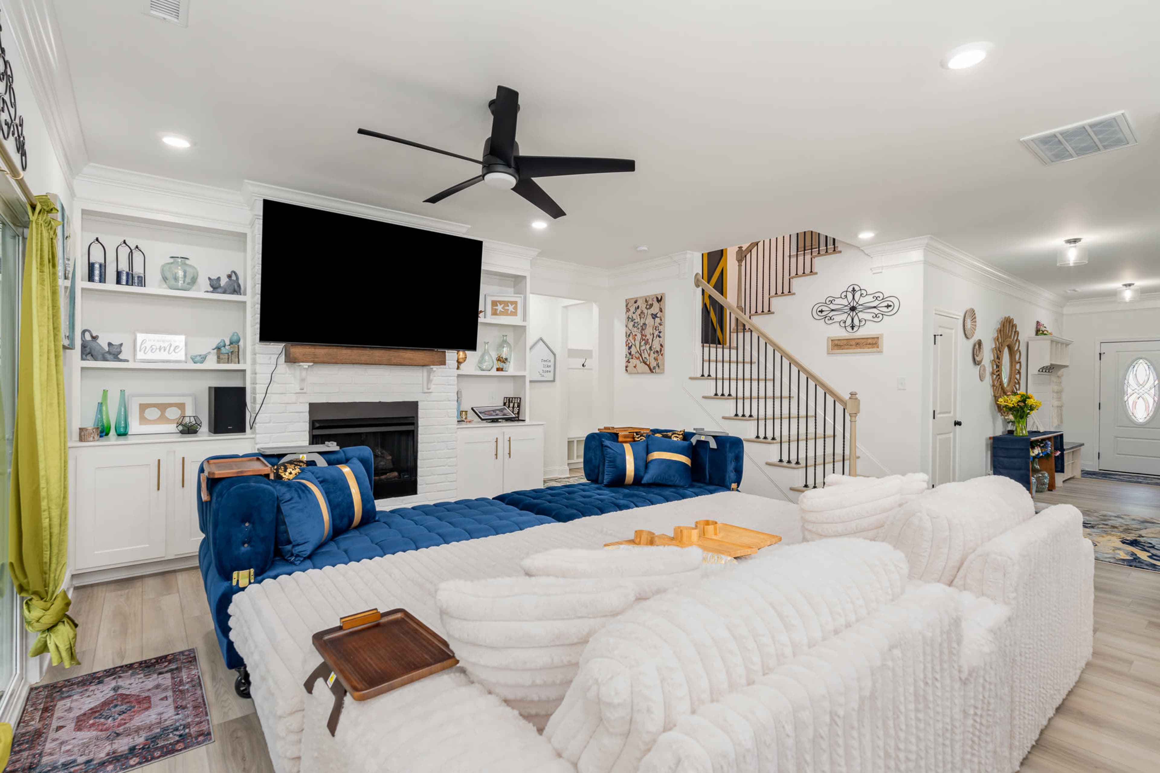 A modern living room features a central seating area with a white sectional sofa and blue accent chairs, a large flat-screen TV above a fireplace, and a staircase in the background.