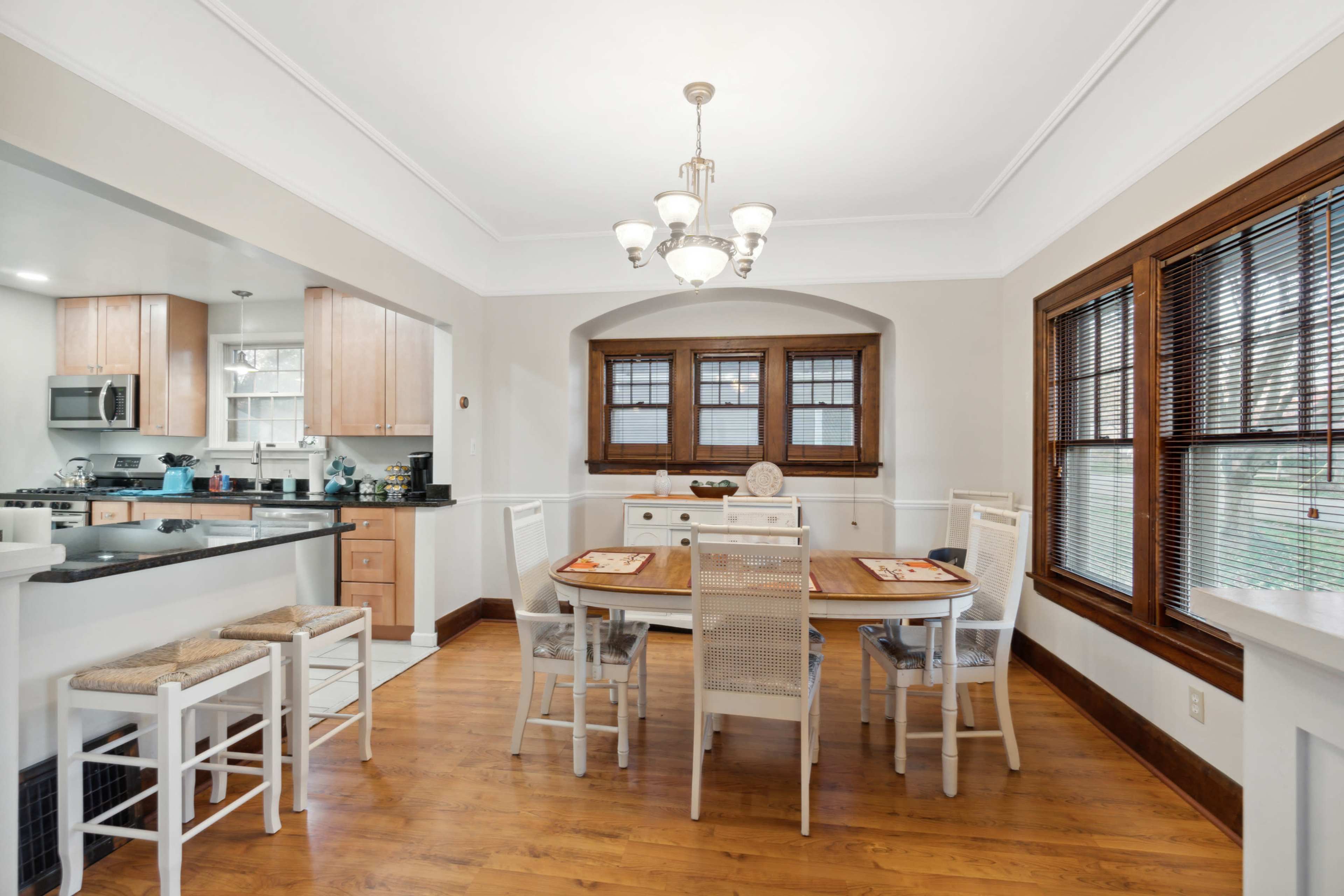 A dining area features a wooden table surrounded by white chairs, with large windows and a chandelier overhead.