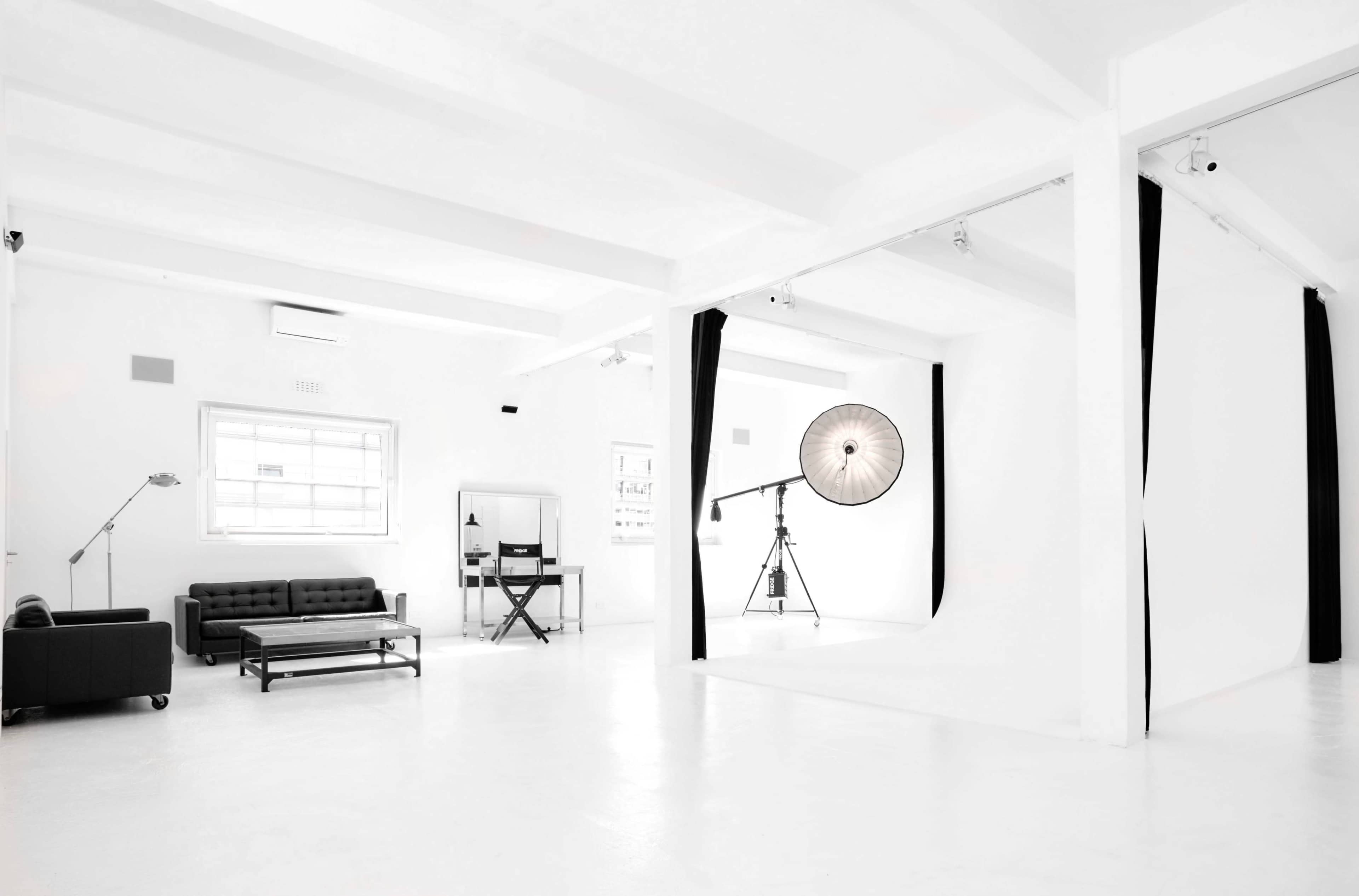 A bright, minimalist studio featuring a black sofa, a director's chair, and a large light setup against a white backdrop.