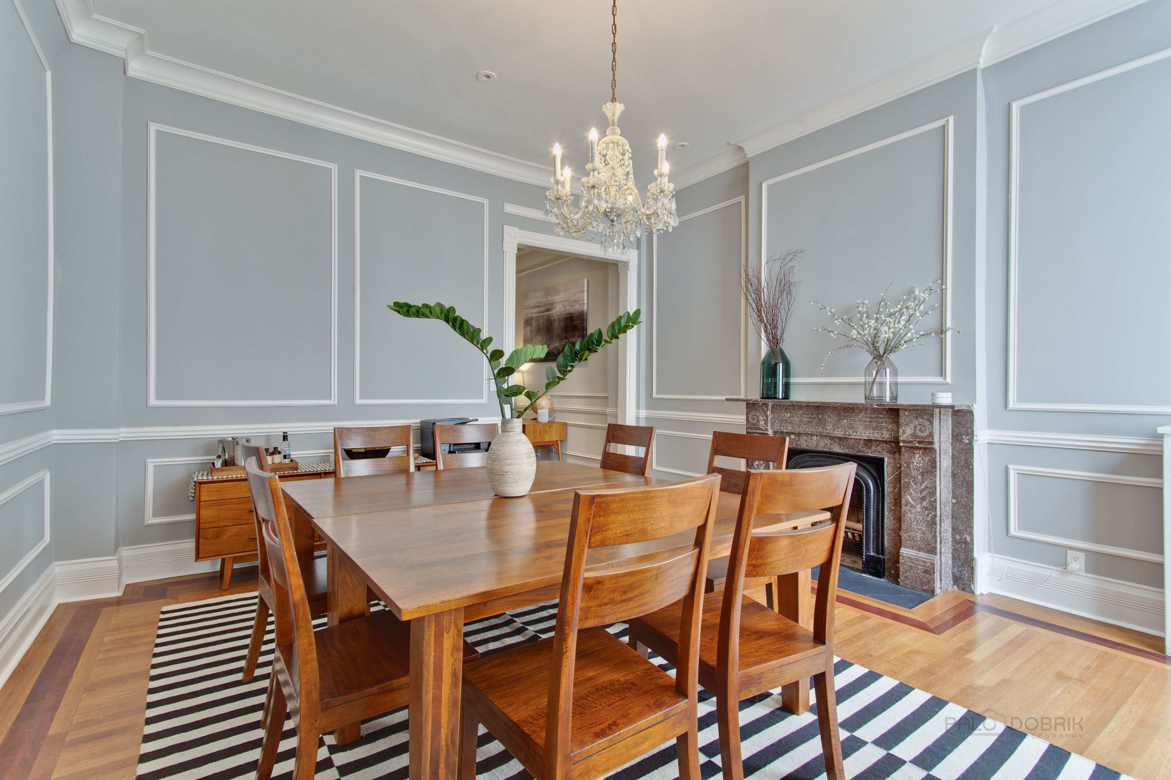 A wooden dining table surrounded by chairs is situated in a room with gray walls and a chandelier, featuring a striped rug and a decorative fireplace.