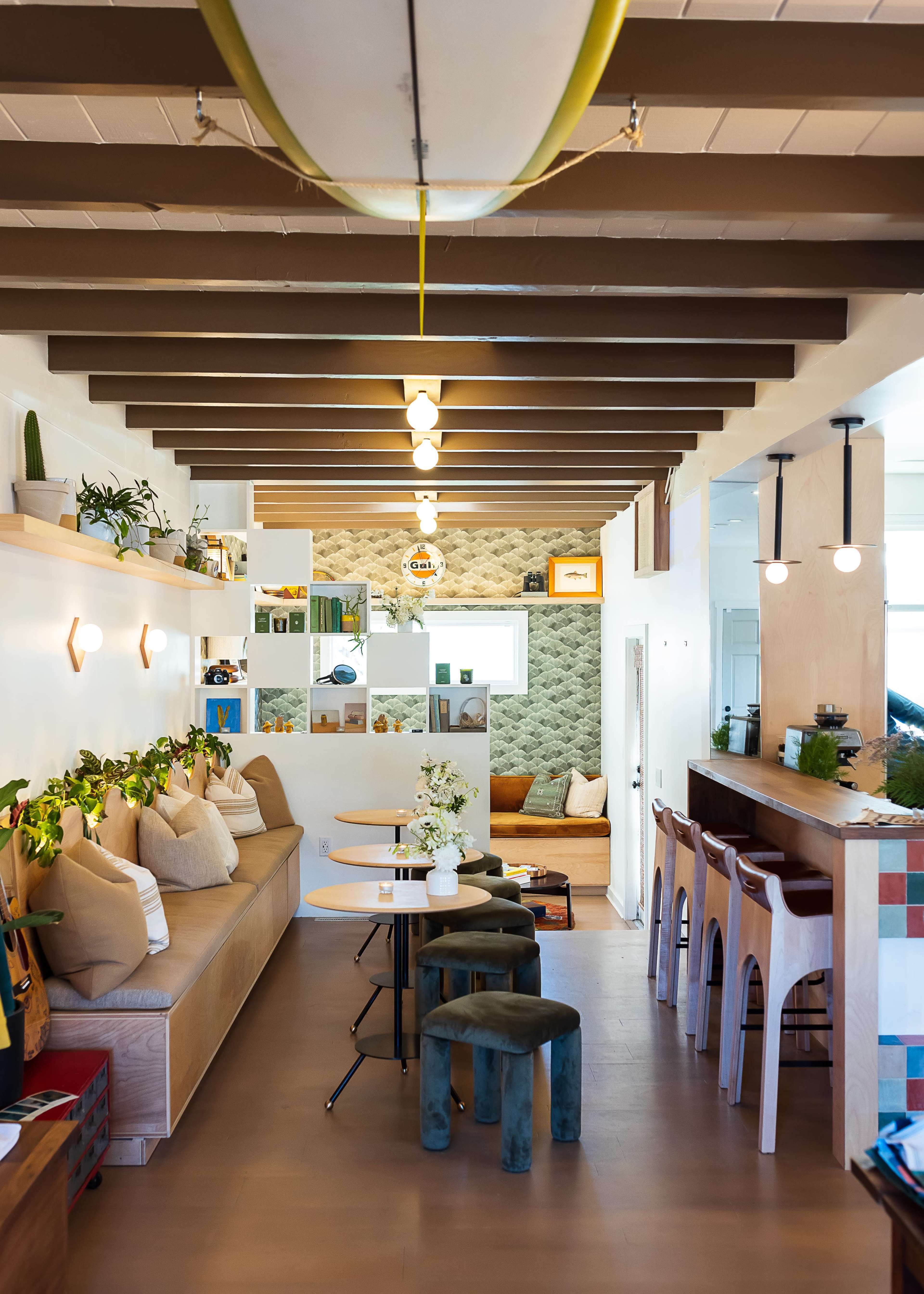 A modern café interior with wooden beams, green accents, seating areas with cushions, and a surfboard hanging from the ceiling.