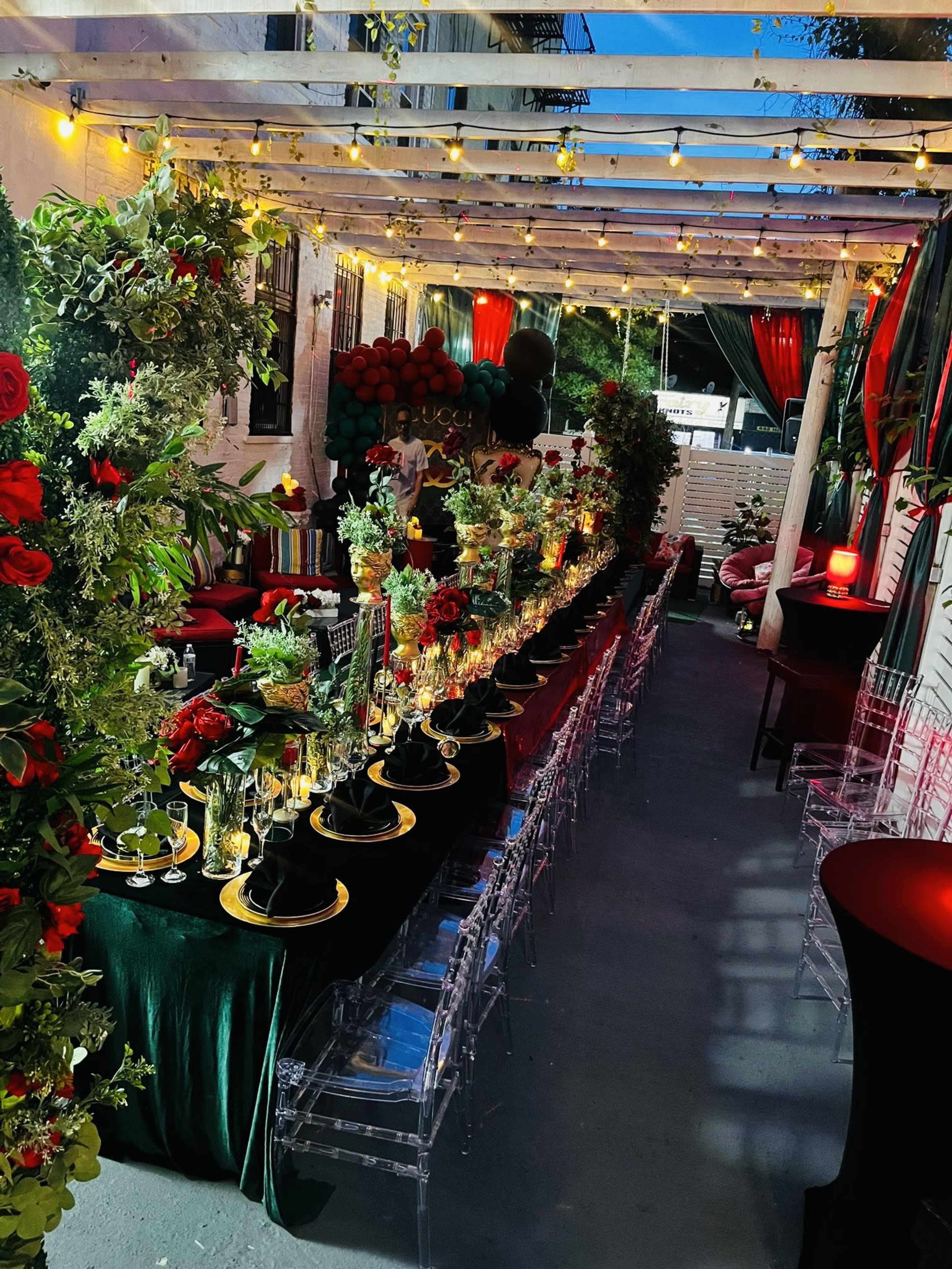 The image shows a decorated outdoor dining area with a long table set for a formal event, featuring floral arrangements, black and gold place settings, and string lights overhead.
