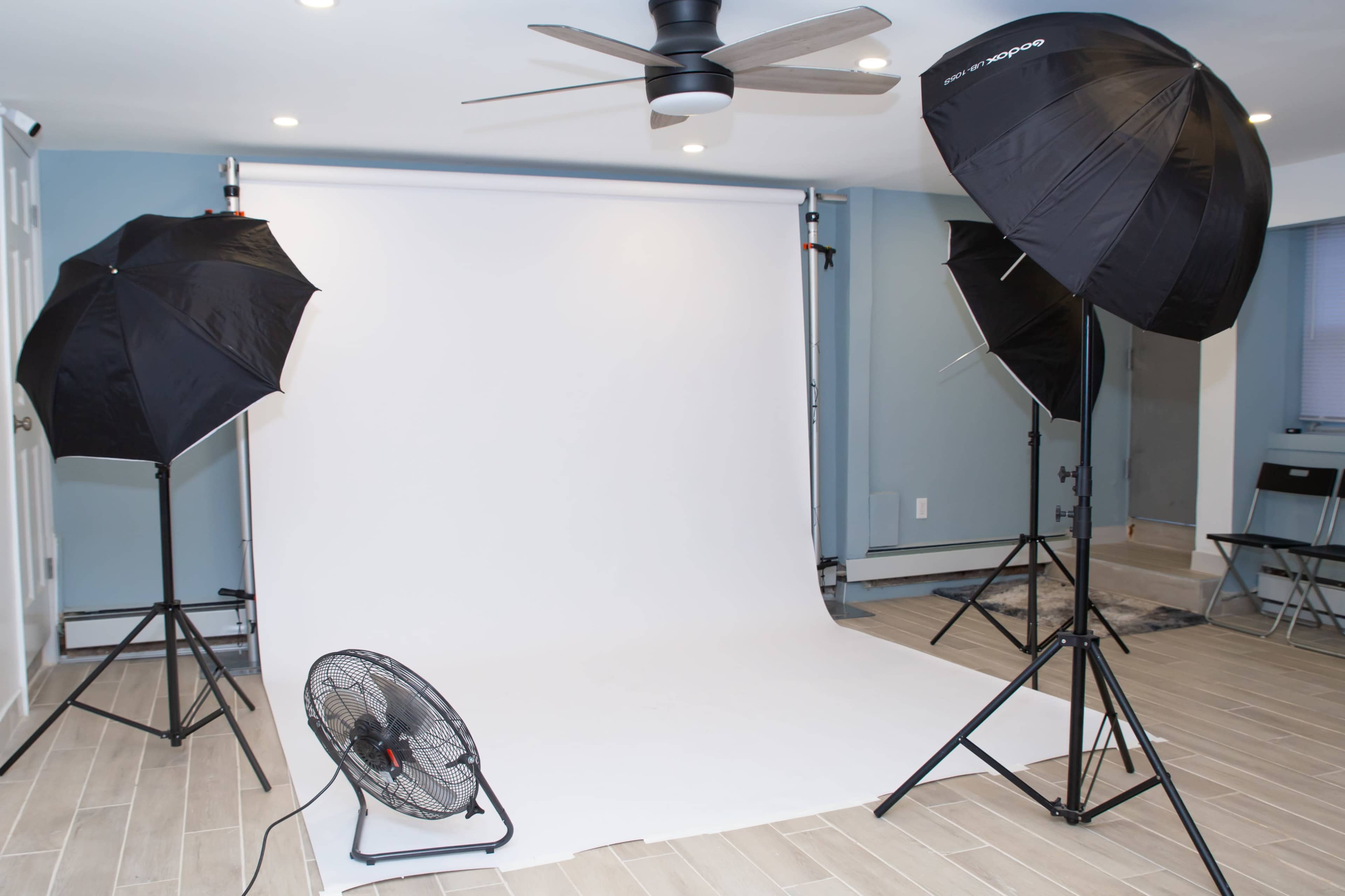 The image shows a photo studio setup with a white backdrop, two softbox lights, and a fan positioned on the floor.
