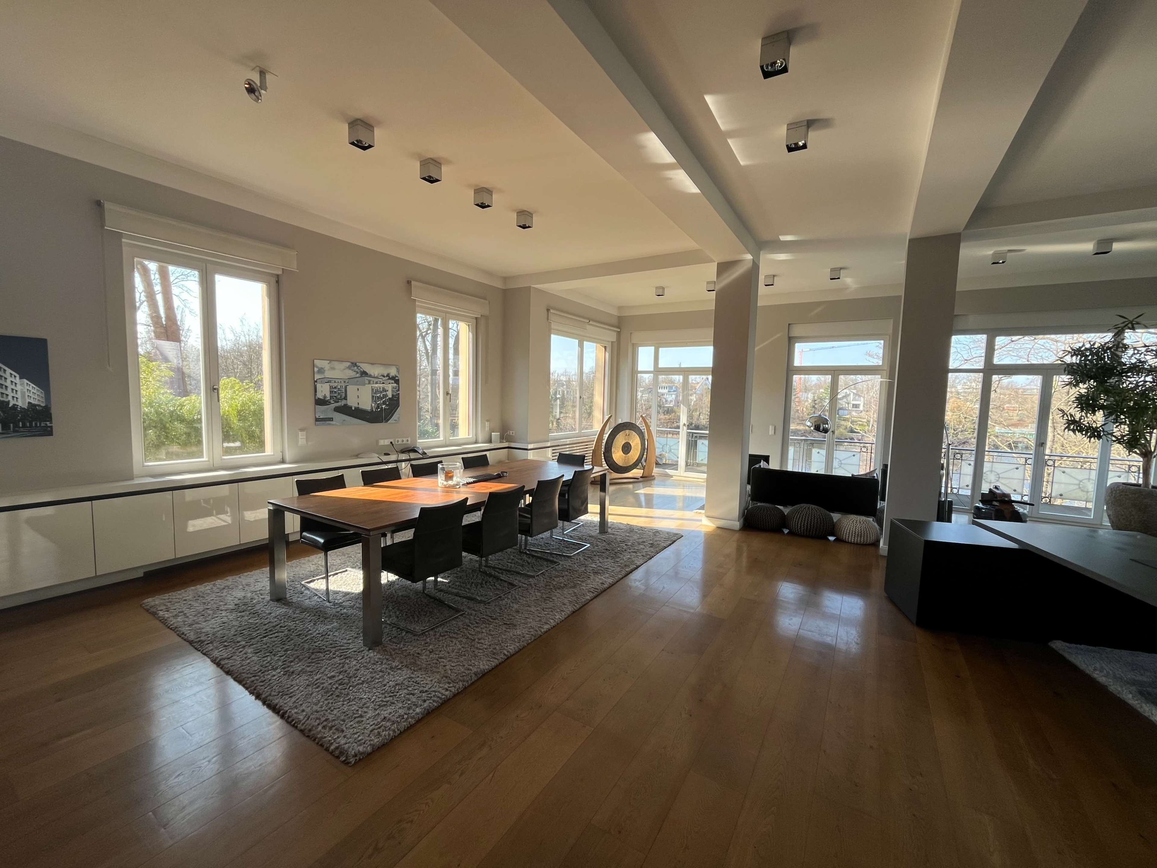 The image shows a spacious living area with a dining table, six chairs, large windows, and a decorative circular feature in the corner.