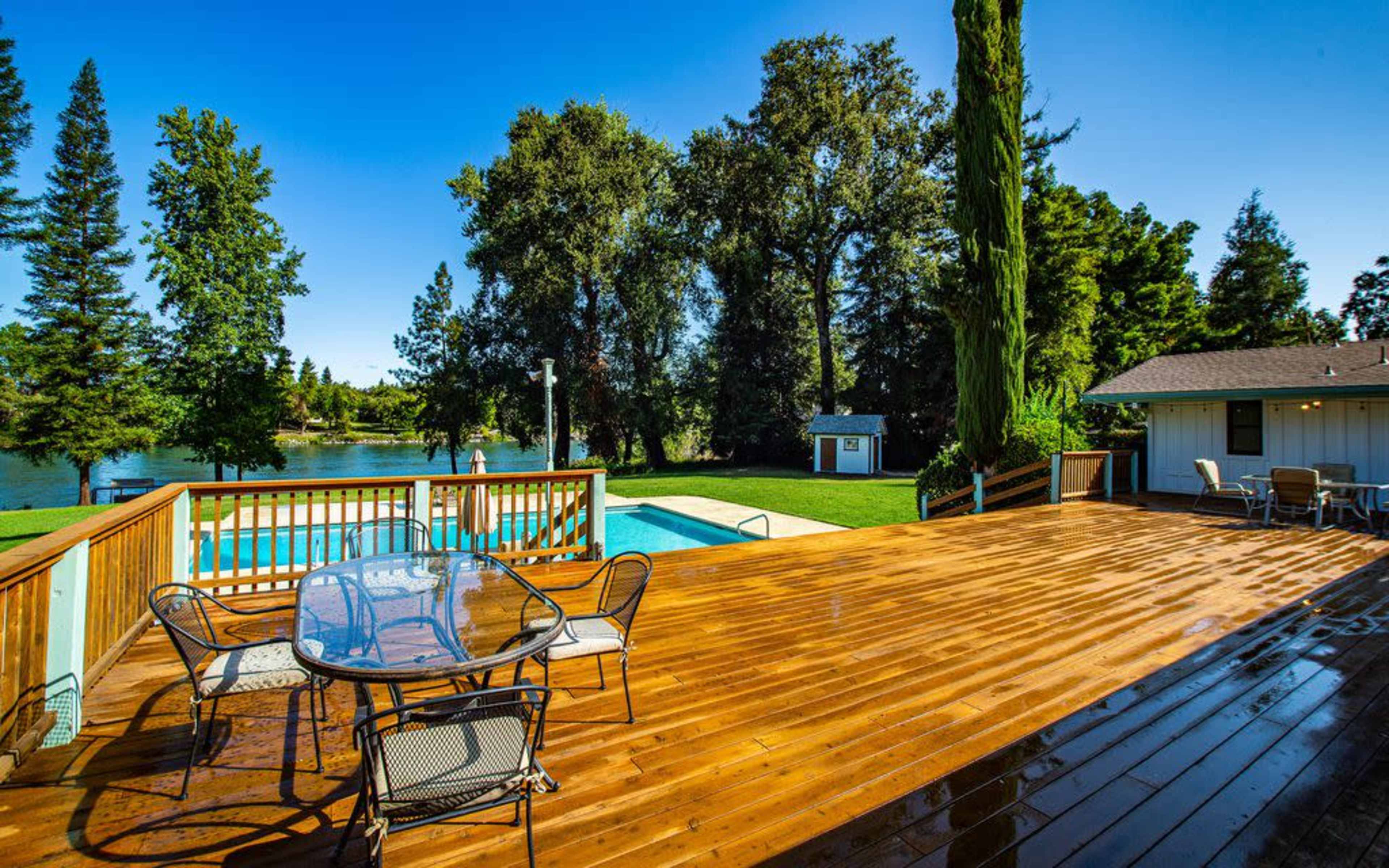 A wooden deck with a glass table and chairs overlooks a swimming pool and a lake surrounded by trees.