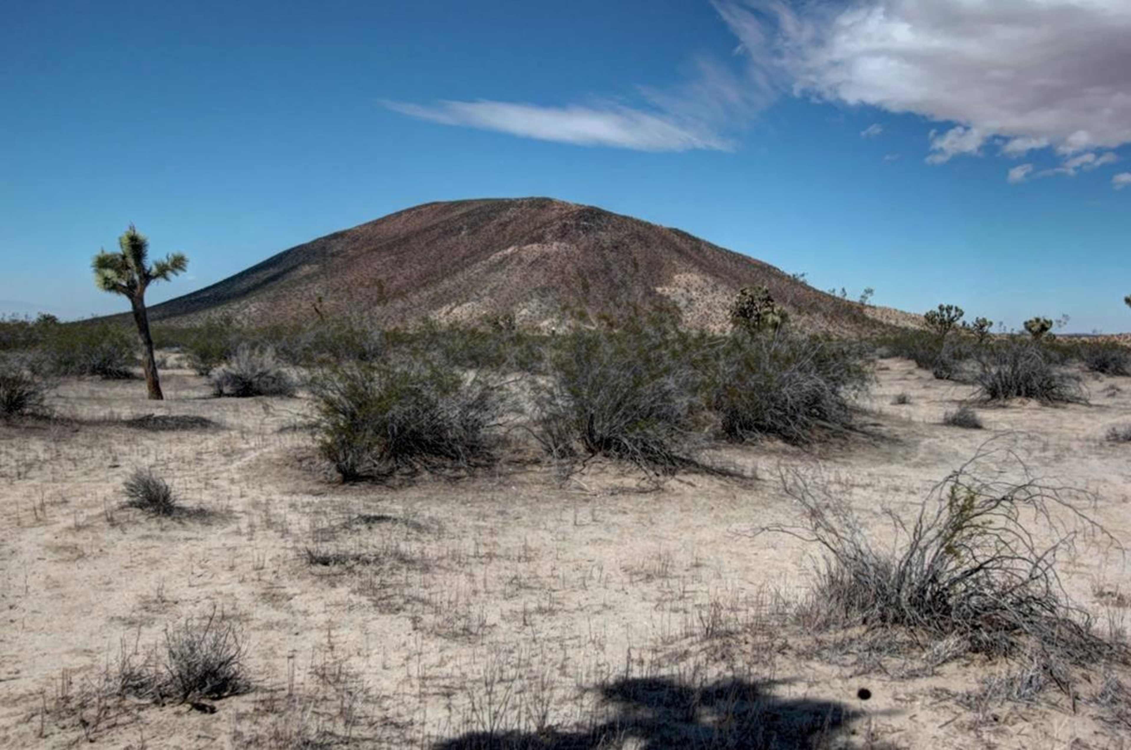 A small, rocky hill rises against a clear blue sky in a arid desert landscape dotted with sparse vegetation.