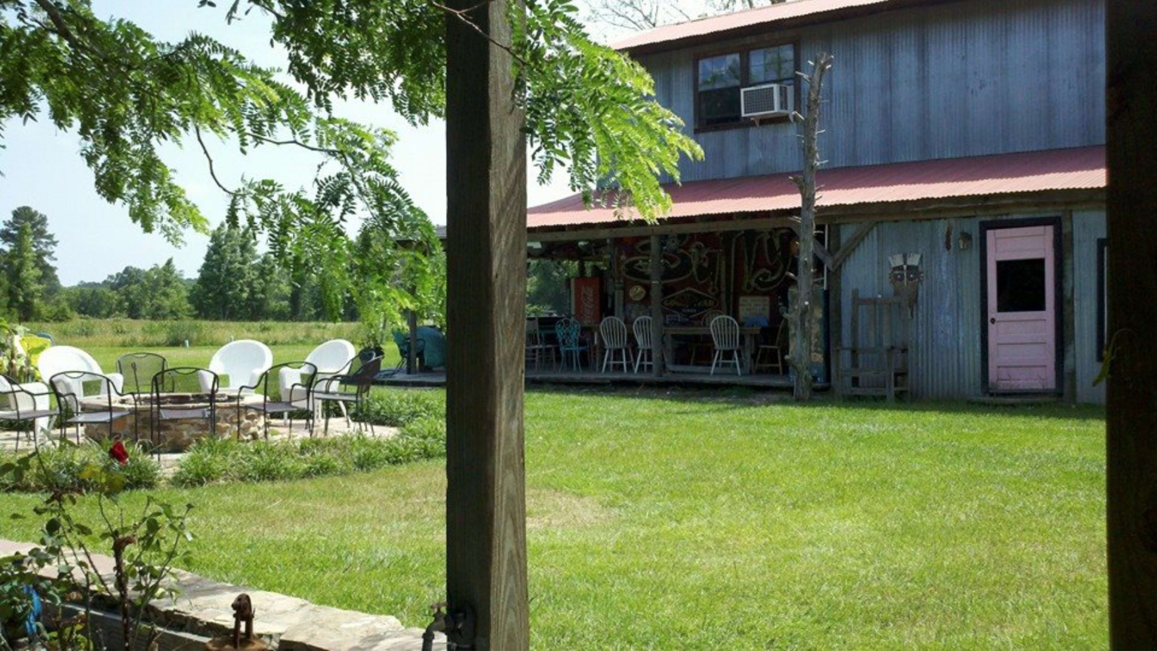 A wooden structure with a red metal roof sits in a grassy area, featuring a fire pit and patio chairs in front.