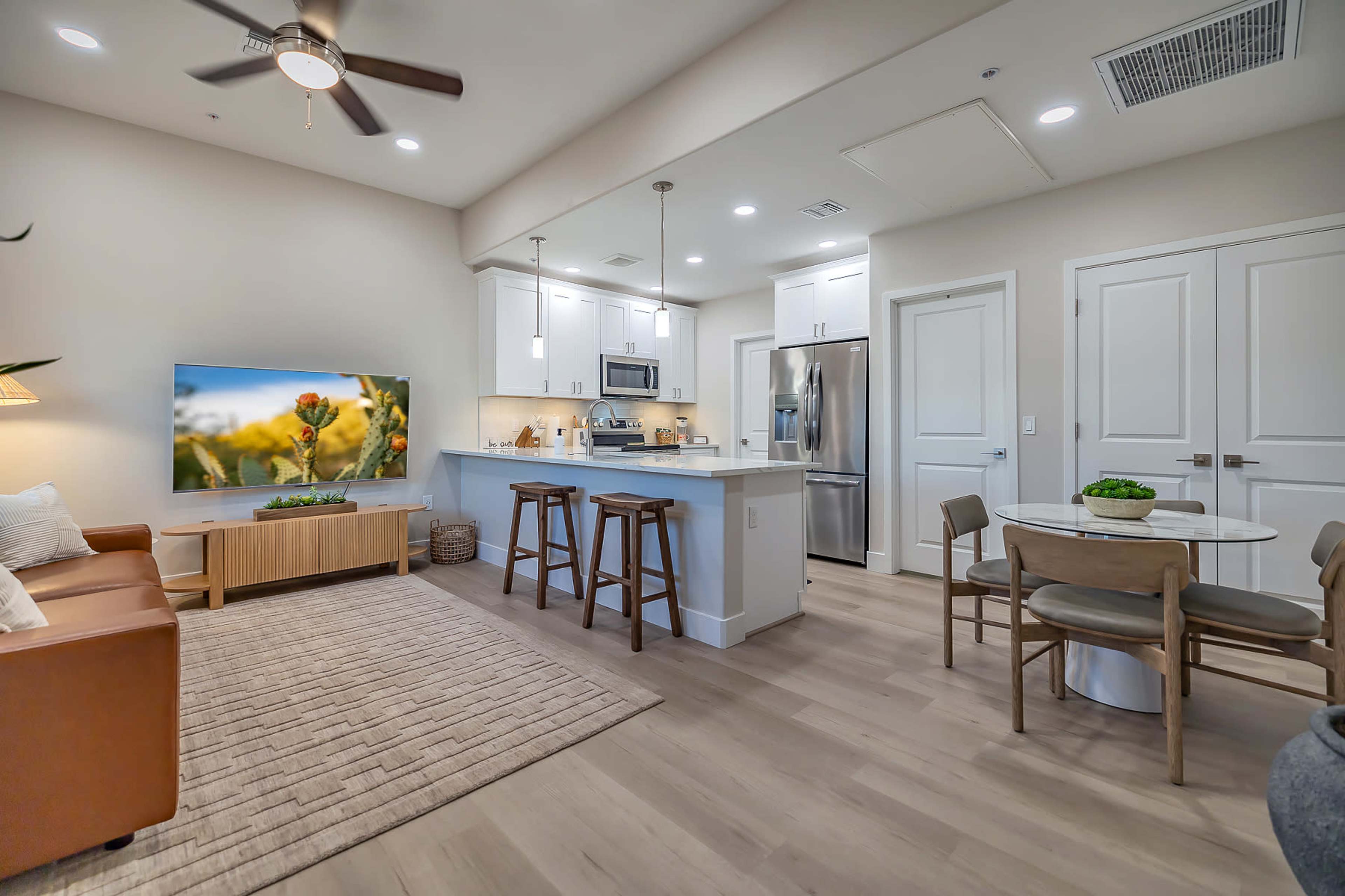 A modern living area with a kitchen, featuring a brown leather sofa, a circular dining table, and a wall-mounted television.