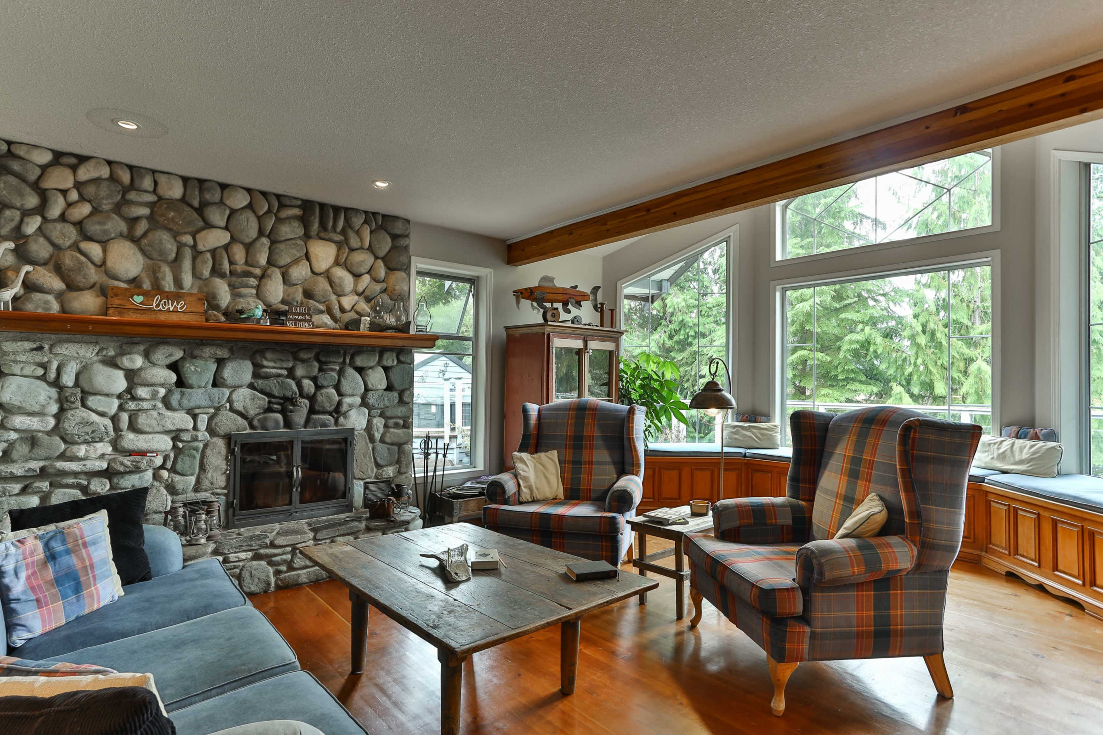 A living room with a stone fireplace, two tufted armchairs, a wooden coffee table, and large windows providing a view of greenery outside.