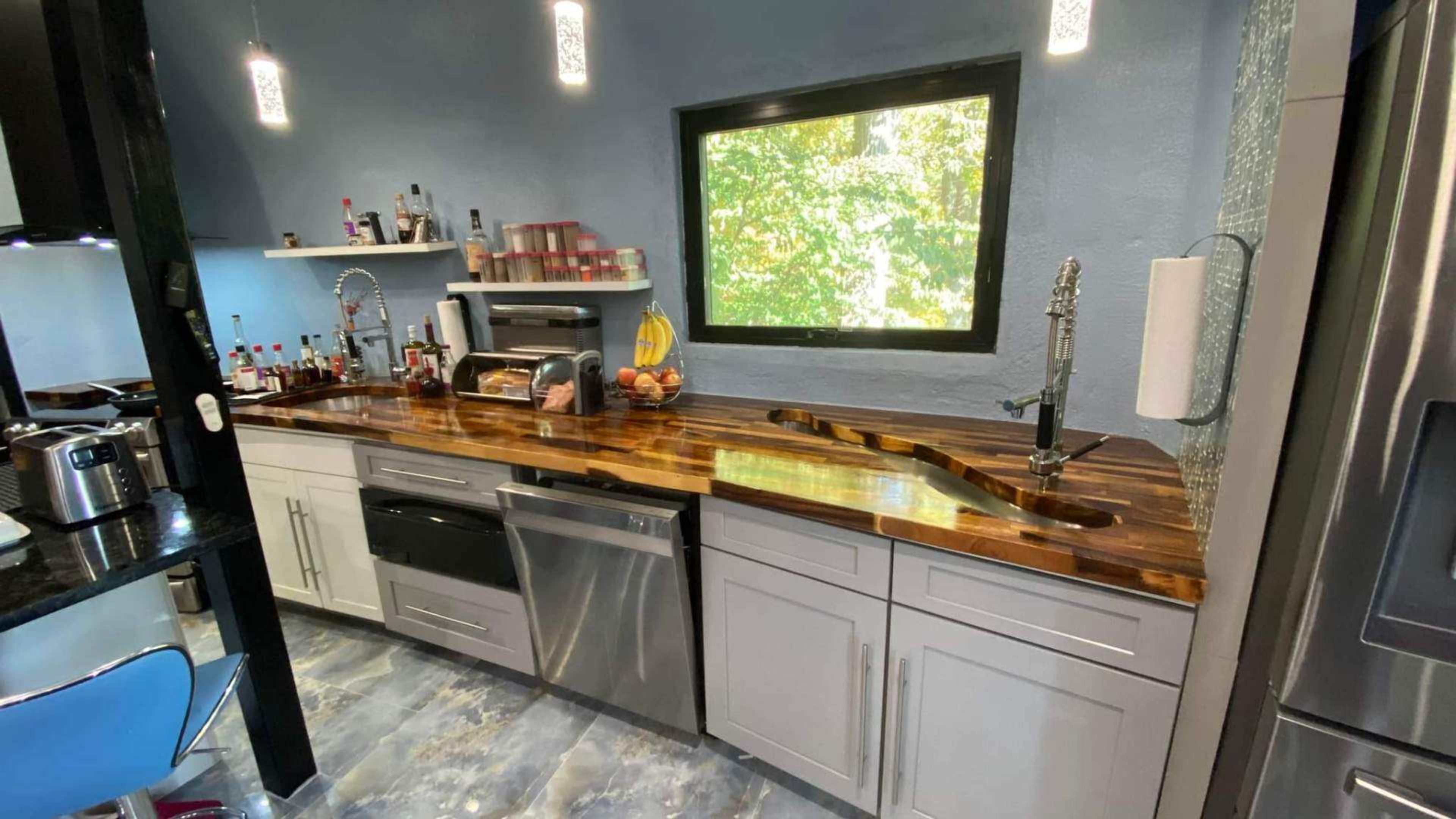 A modern kitchen with a wooden countertop, stainless steel appliances, and a large window displaying a green outdoor view.