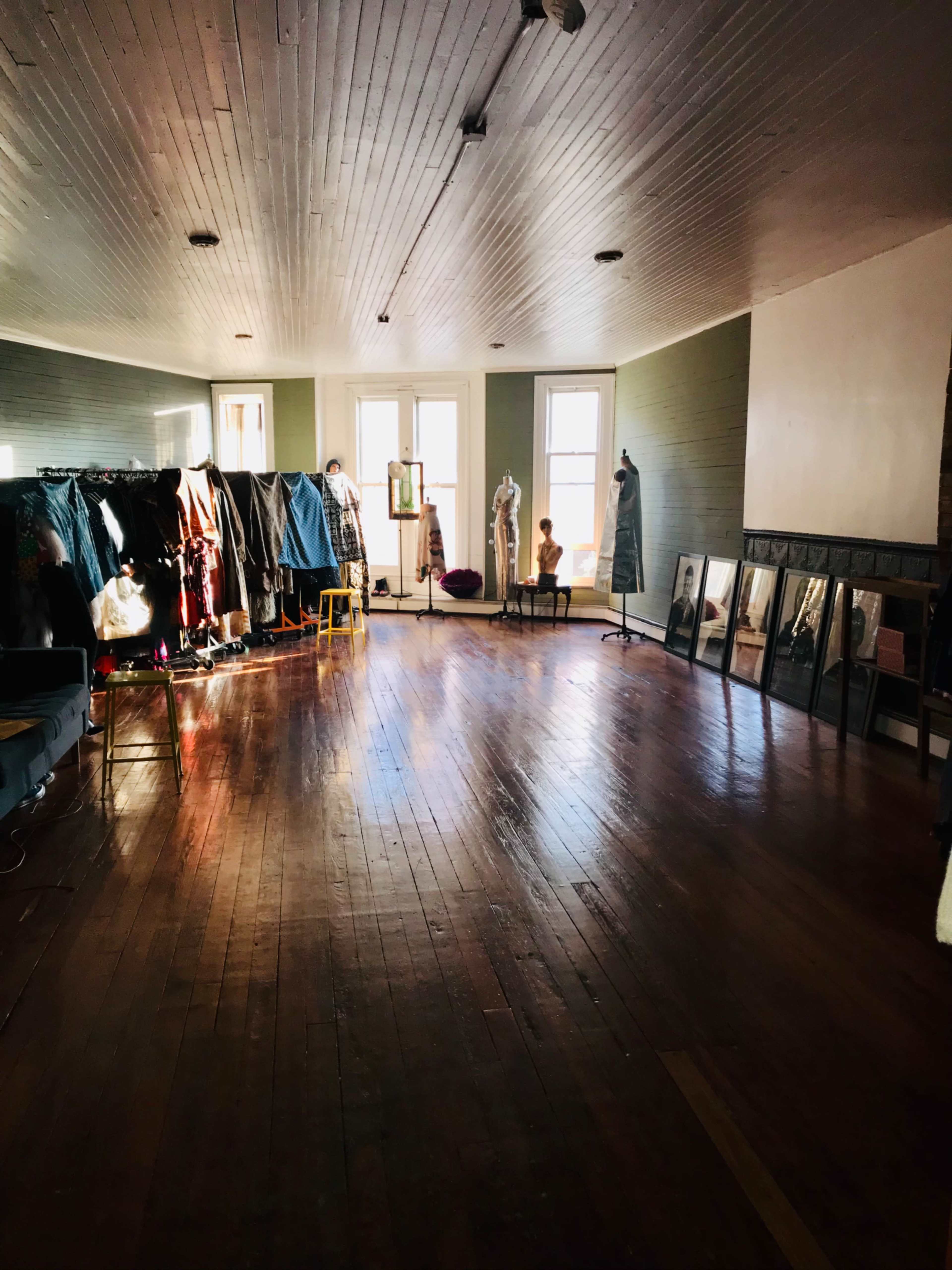 The image shows a spacious room with wooden floors, featuring racks of clothing along one wall and large windows allowing natural light to illuminate the space.