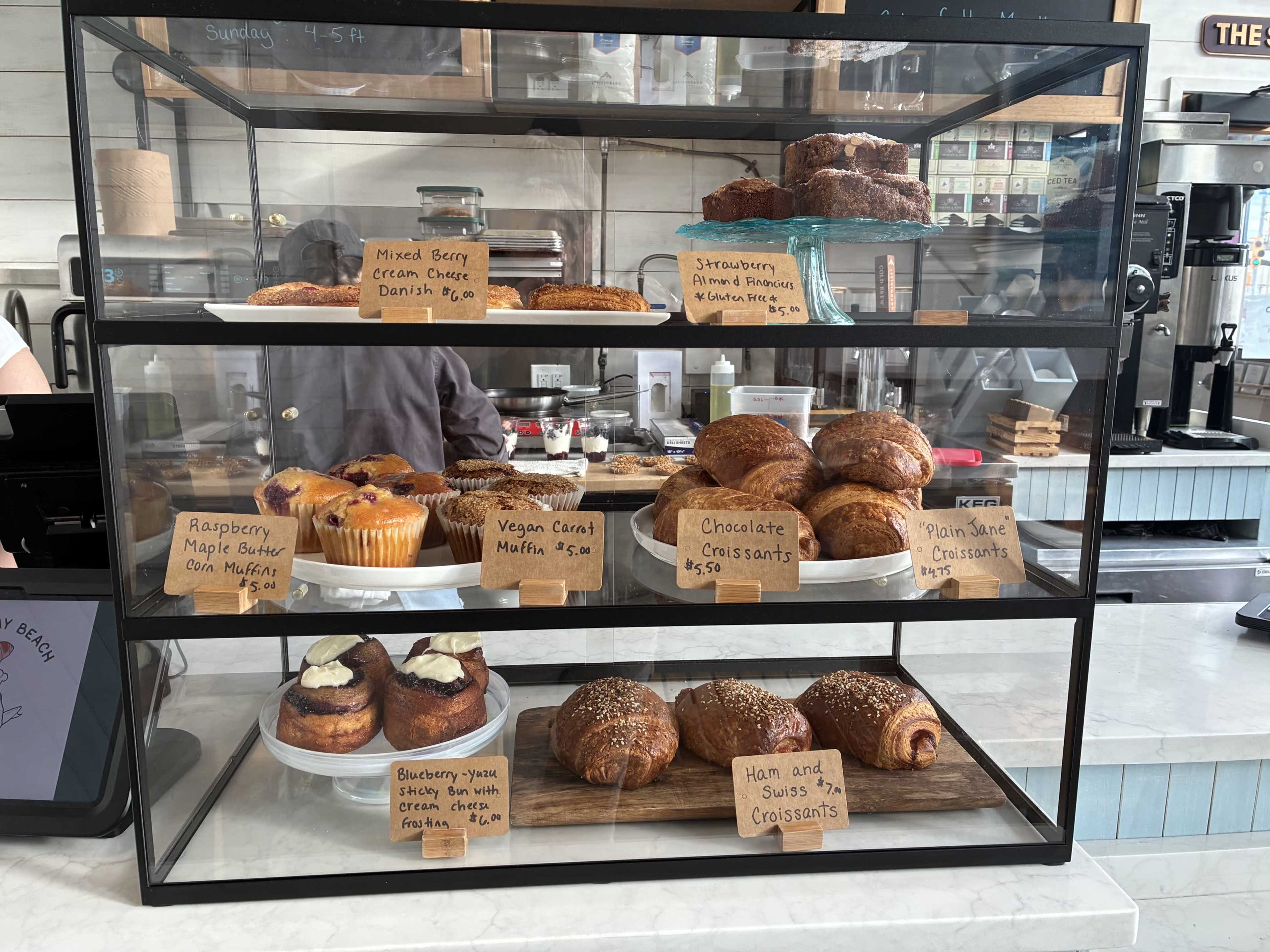 The display case contains various baked goods, including muffins, croissants, and danishes, each labeled with prices.
