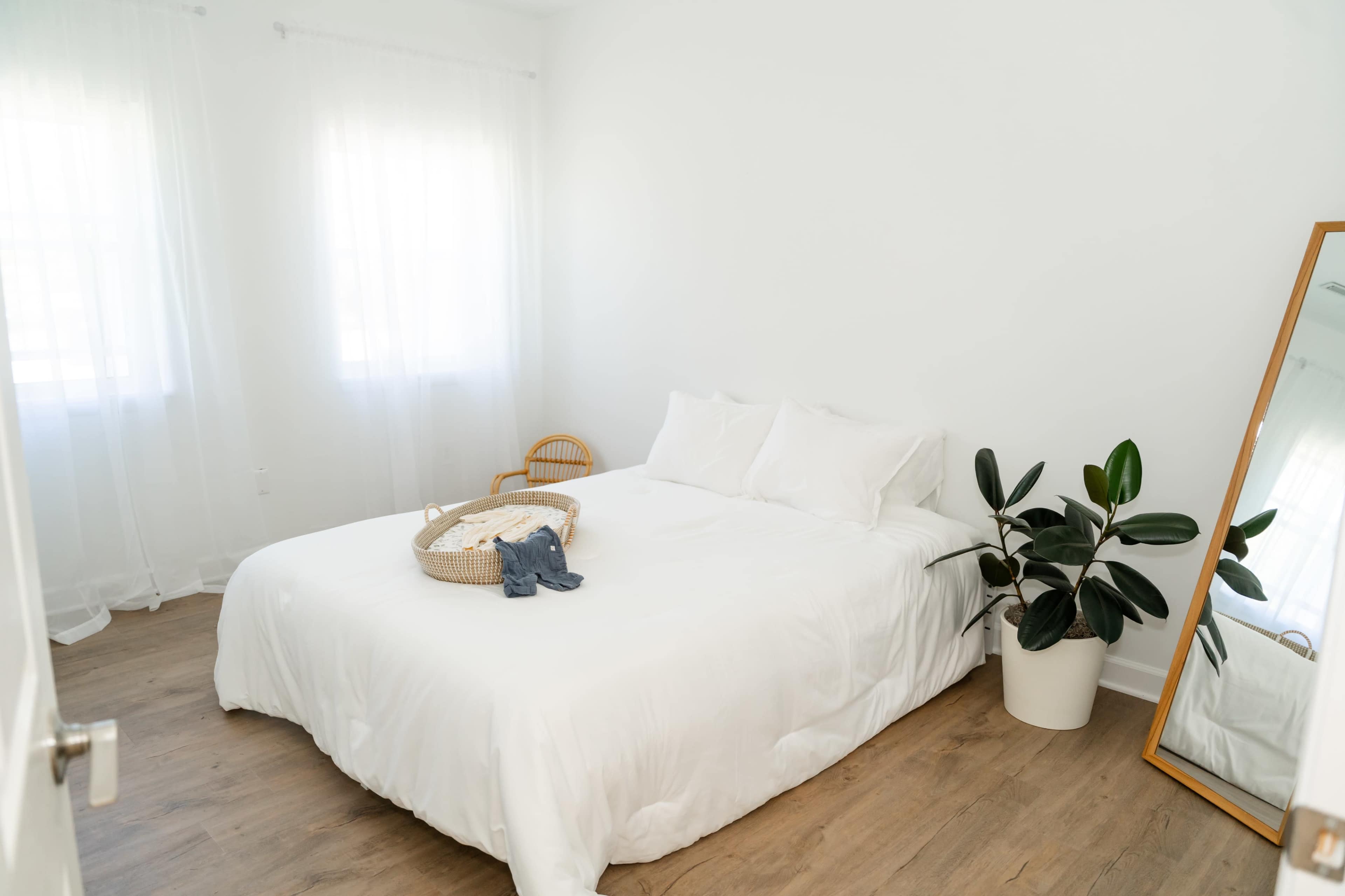 The image shows a minimalistic bedroom featuring a large white bed with a woven basket on top, a potted plant, and a full-length mirror against a white wall.