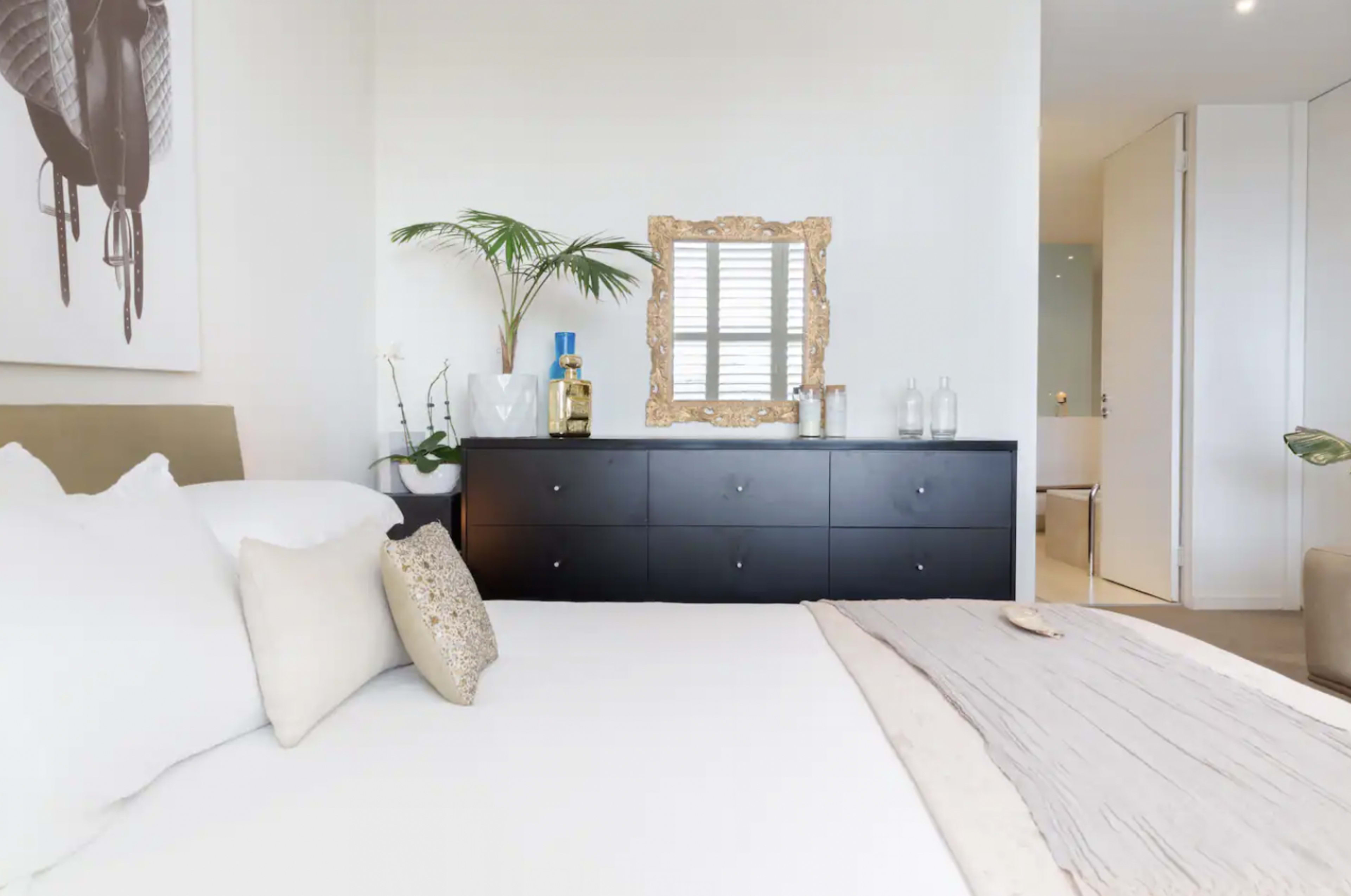 A modern bedroom featuring a bed with white linens, a black dresser, a decorative mirror, and a potted plant.