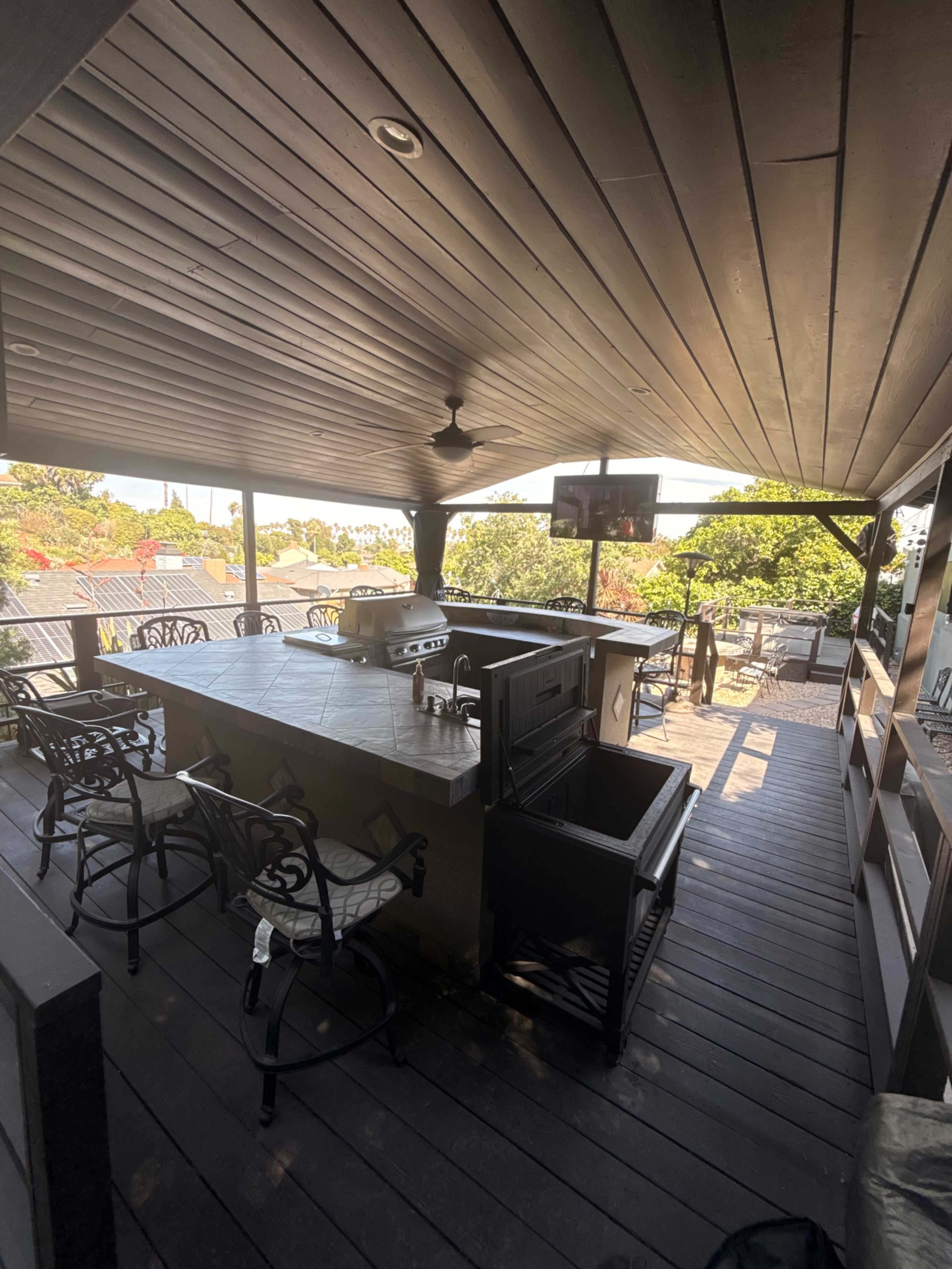 The image shows a spacious outdoor kitchen area with a grill, bar seating, and a mounted television under a wooden covered patio.