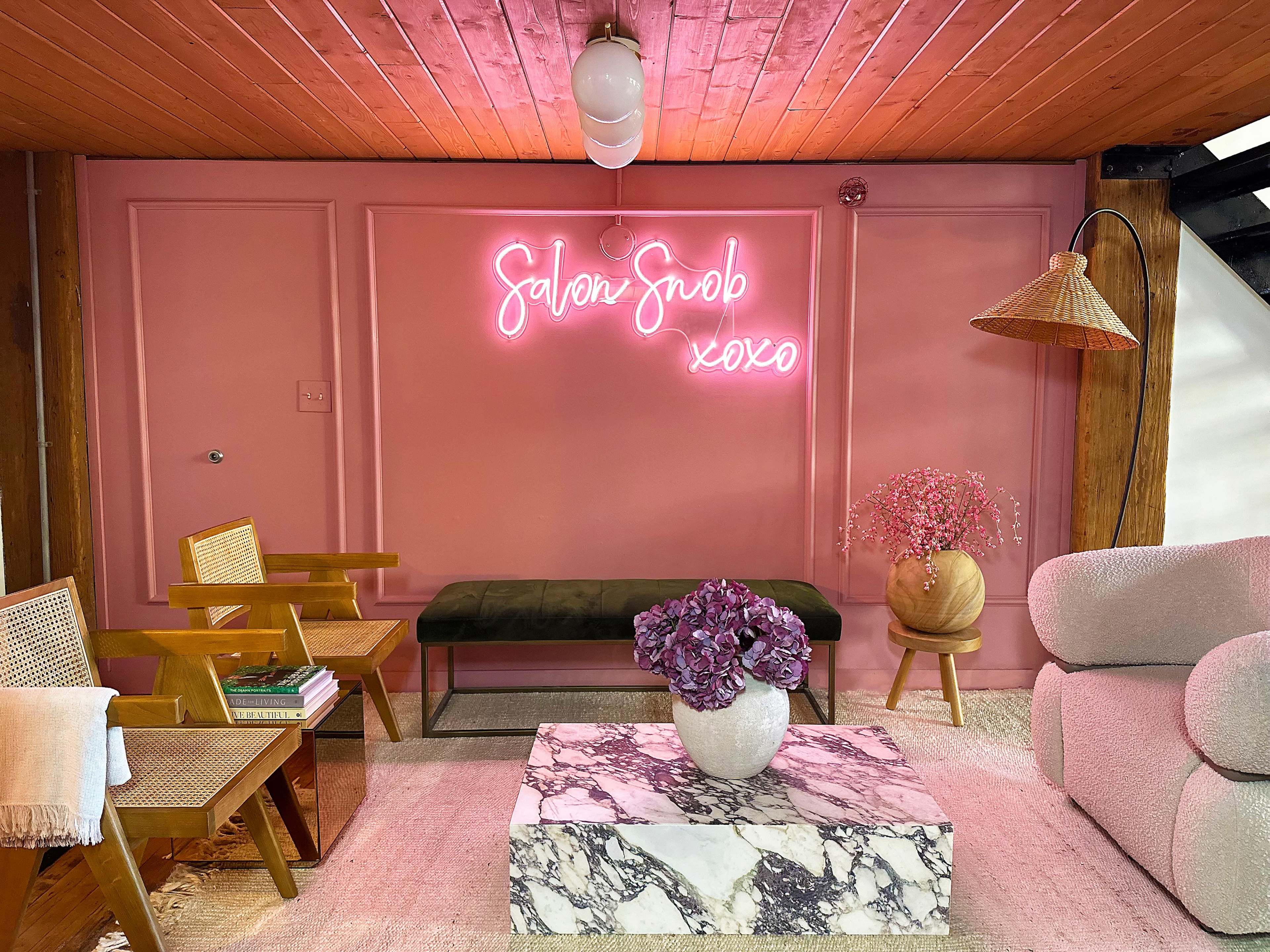 A stylish interior with a pink wall, a neon sign reading "Salon Snob xoxo," a marble coffee table, and wooden chairs.
