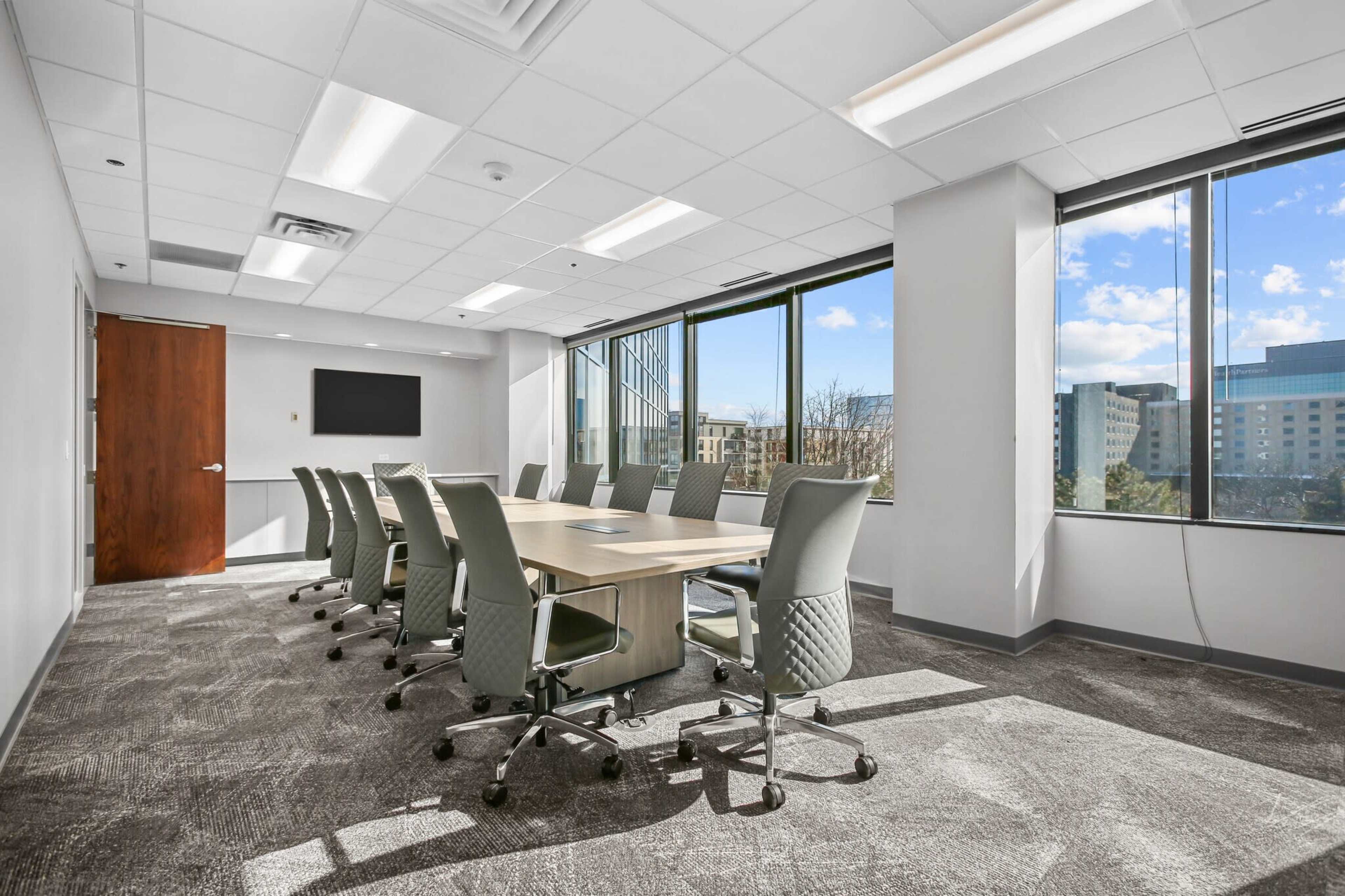 A well-lit conference room features a large table surrounded by ten ergonomic chairs and large windows offering a view of the outside landscape.