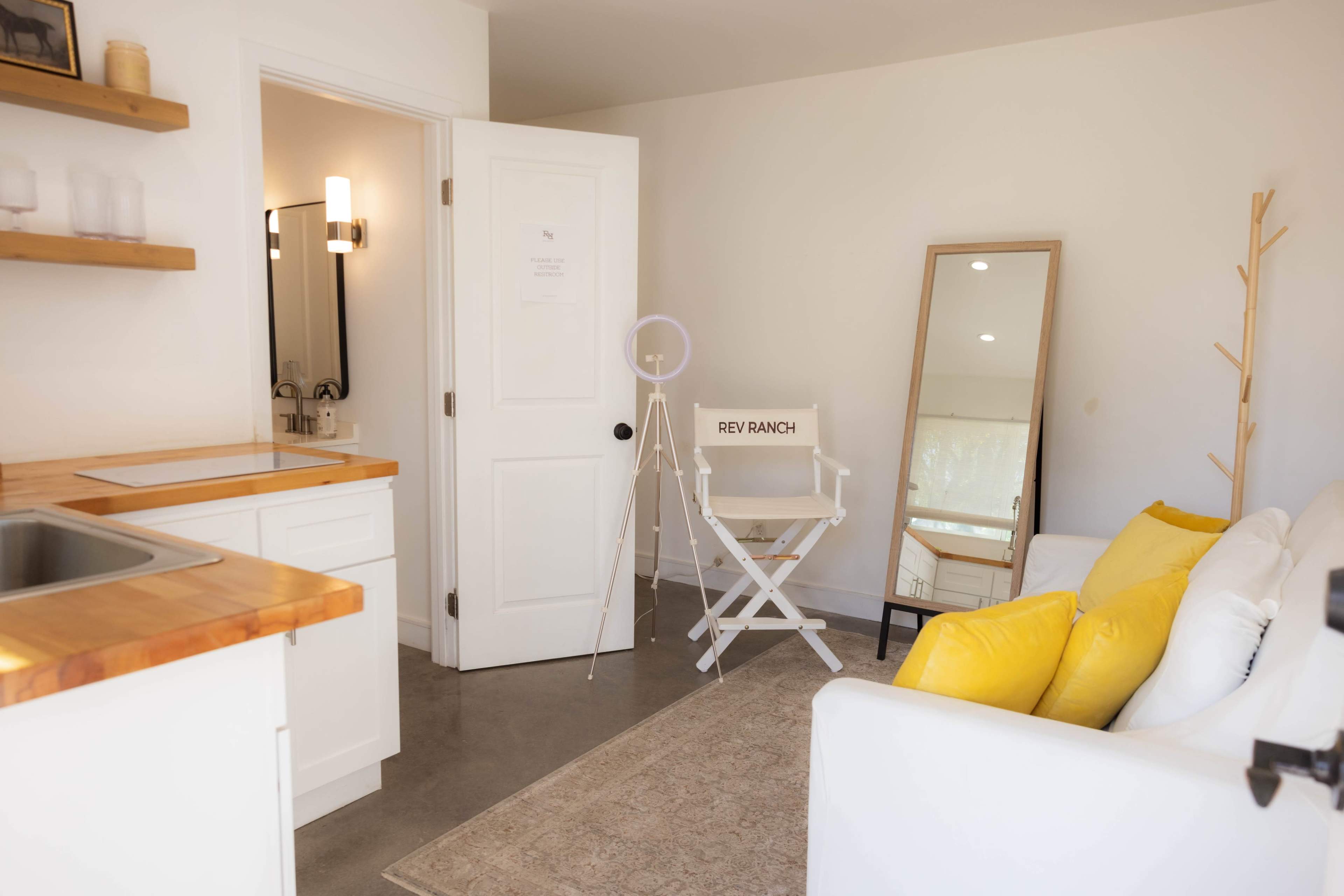 The image shows a minimalist room with a small kitchen area, a mirror, a sitting area with a white sofa and yellow cushions, and a director's chair labeled "REY RANCH."